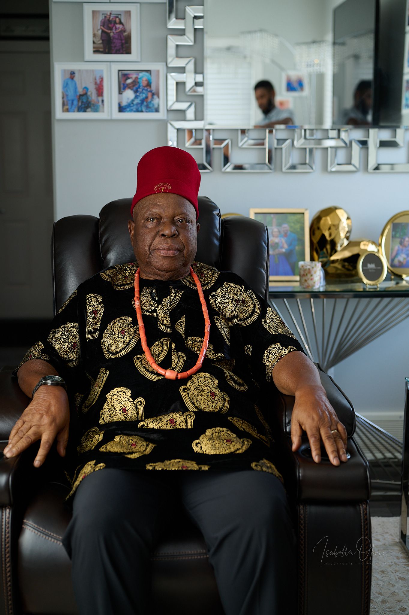 Elder in traditional Nigerian attire with red cap seated in decorated room.