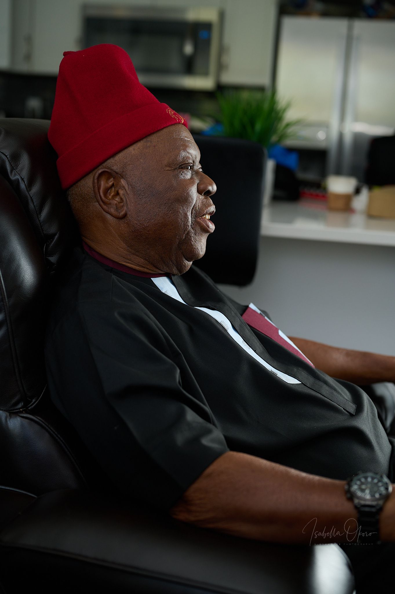 Elder wearing traditional red cap seated in leather chair during formal occasion.