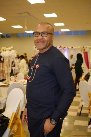 Man in navy blue suit and red beaded necklace at formal event.