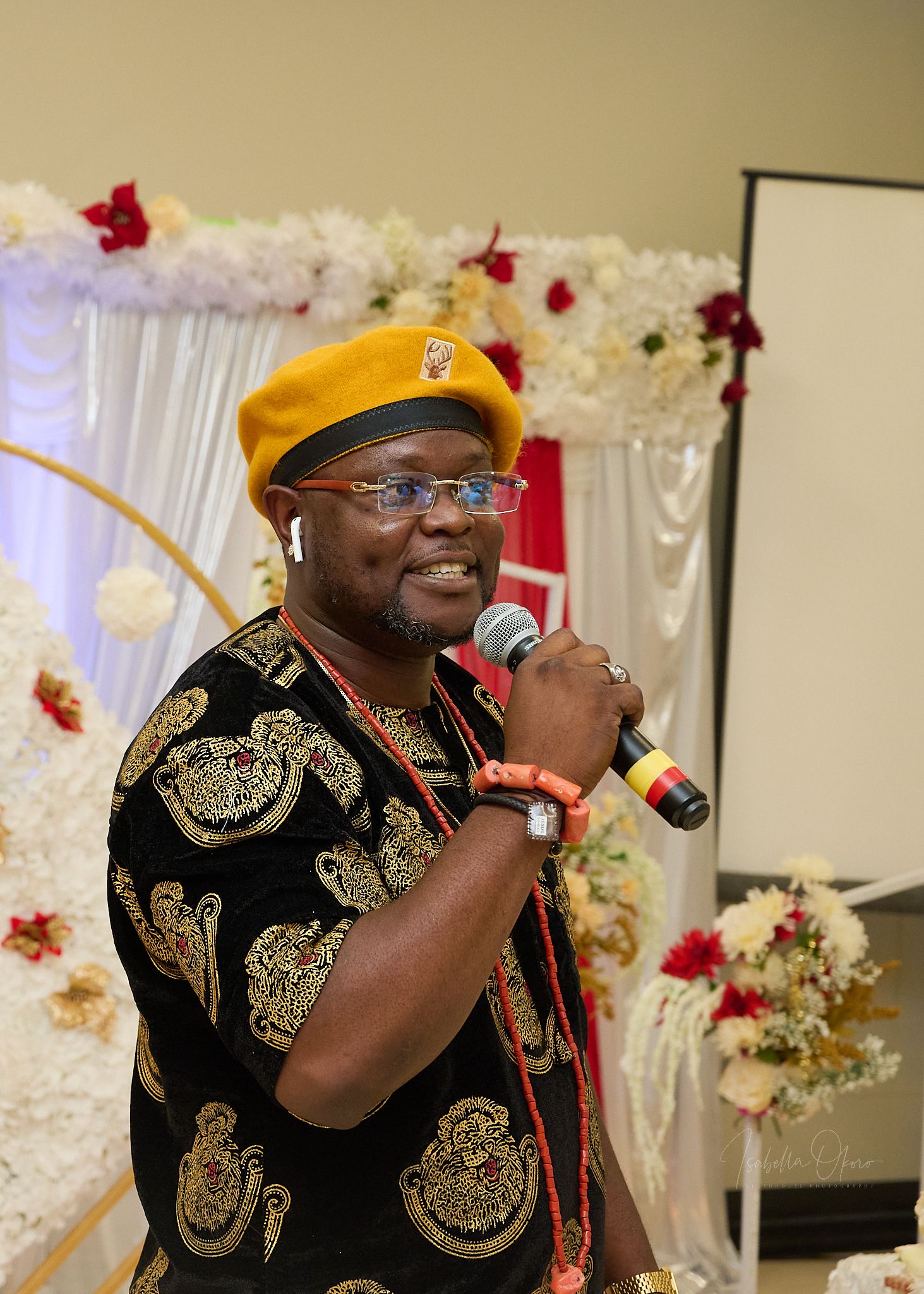 Speaker wearing traditional black and gold patterned attire with yellow cap performs at decorated event.