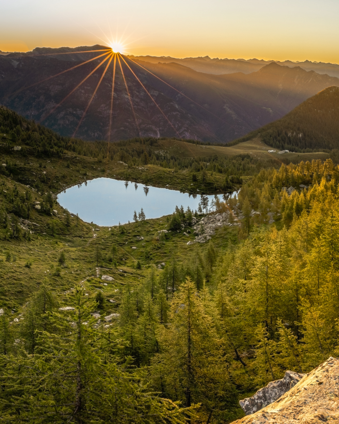 Mountain landscape at sunset with sunburst over peaks reflecting in an alpine lake surrounded by autumn larches.