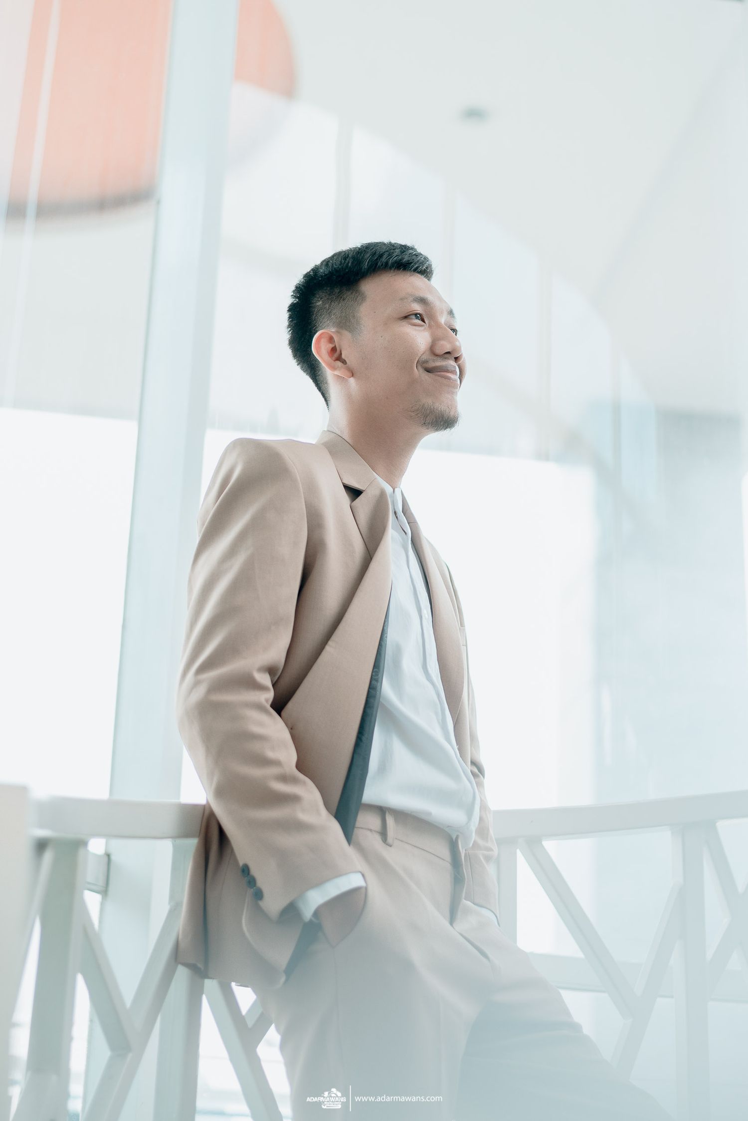 A person in a beige suit jacket smiles while standing in a bright modern interior.