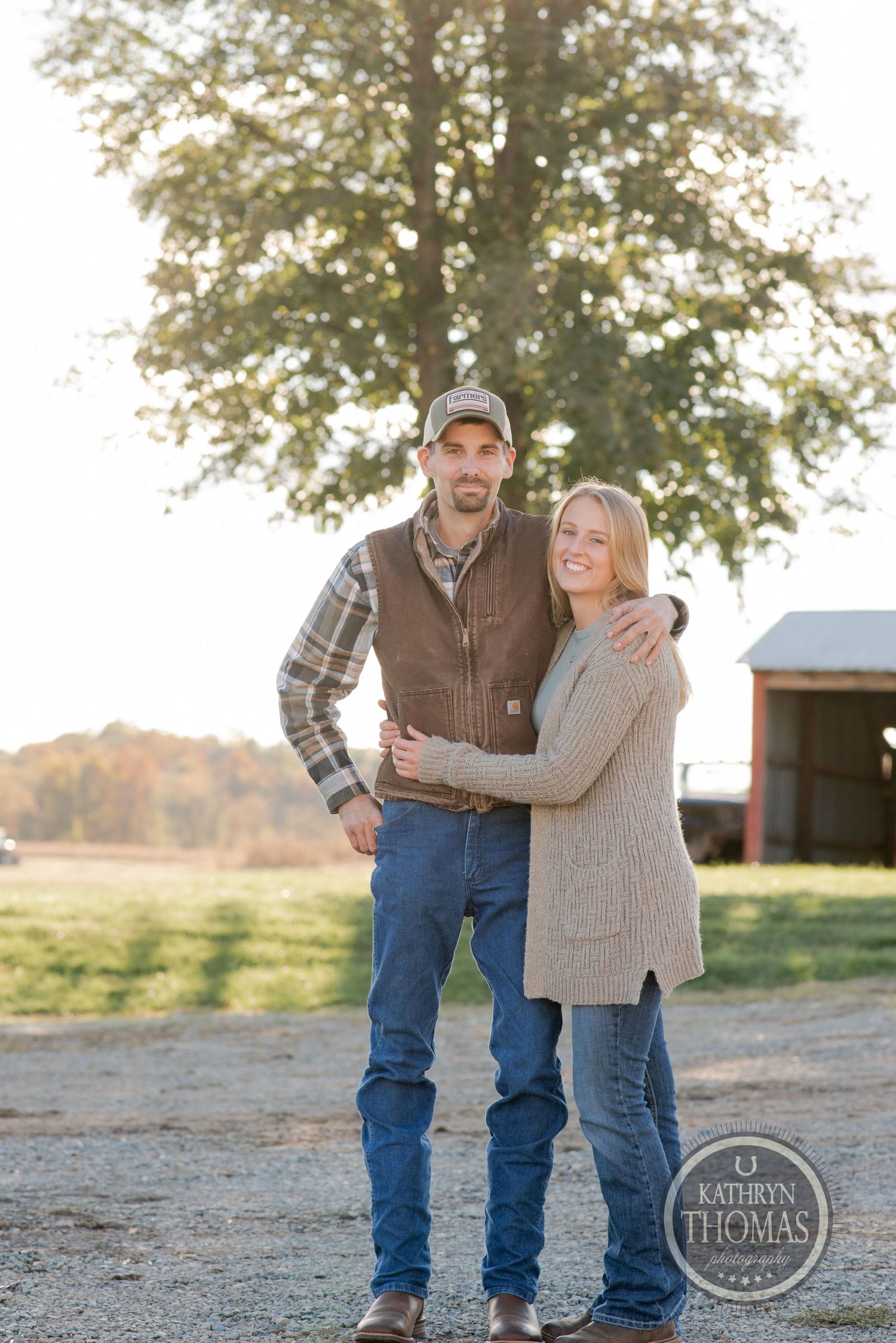 Couple standing together on farm path with autumn trees and barn in background.
