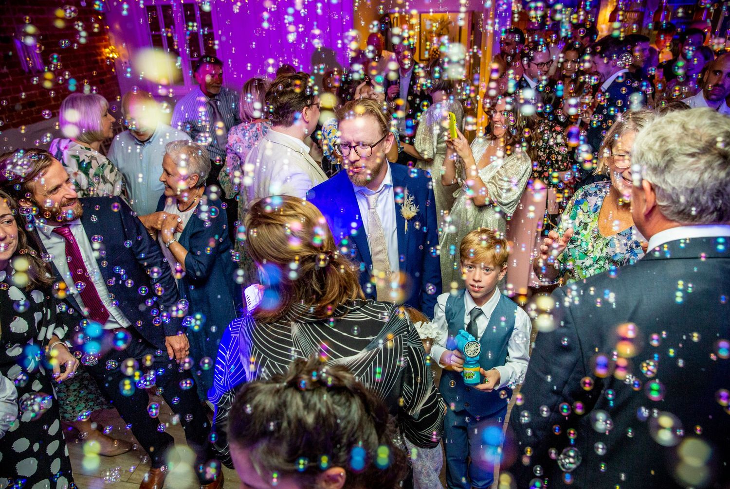 Wedding celebration with guests dancing amid a shower of bubbles under purple lighting.