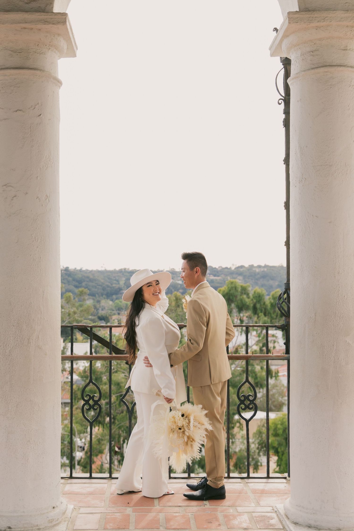 Elegant couple in wedding attire standing between white columns on a balcony overlooking nature.