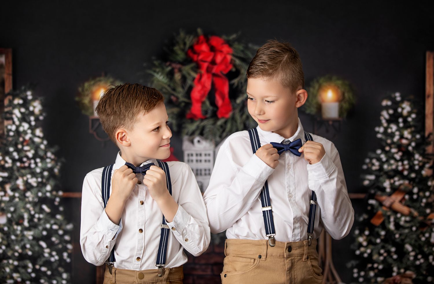 Two children in white shirts and suspenders adjust their bowties against a dark Christmas backdrop with red bows.