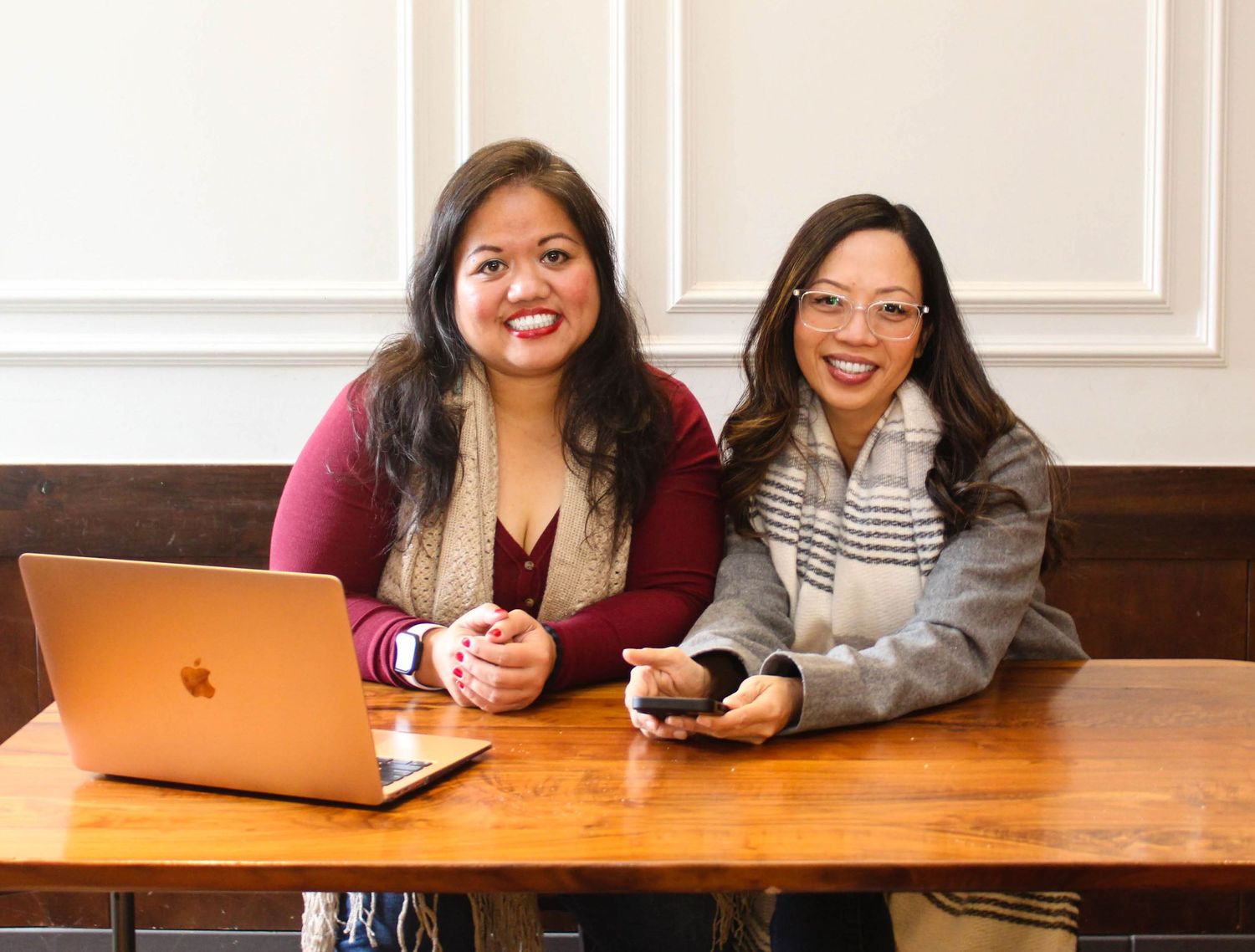 Two friends sitting at wooden table with laptop and phones in casual setting.