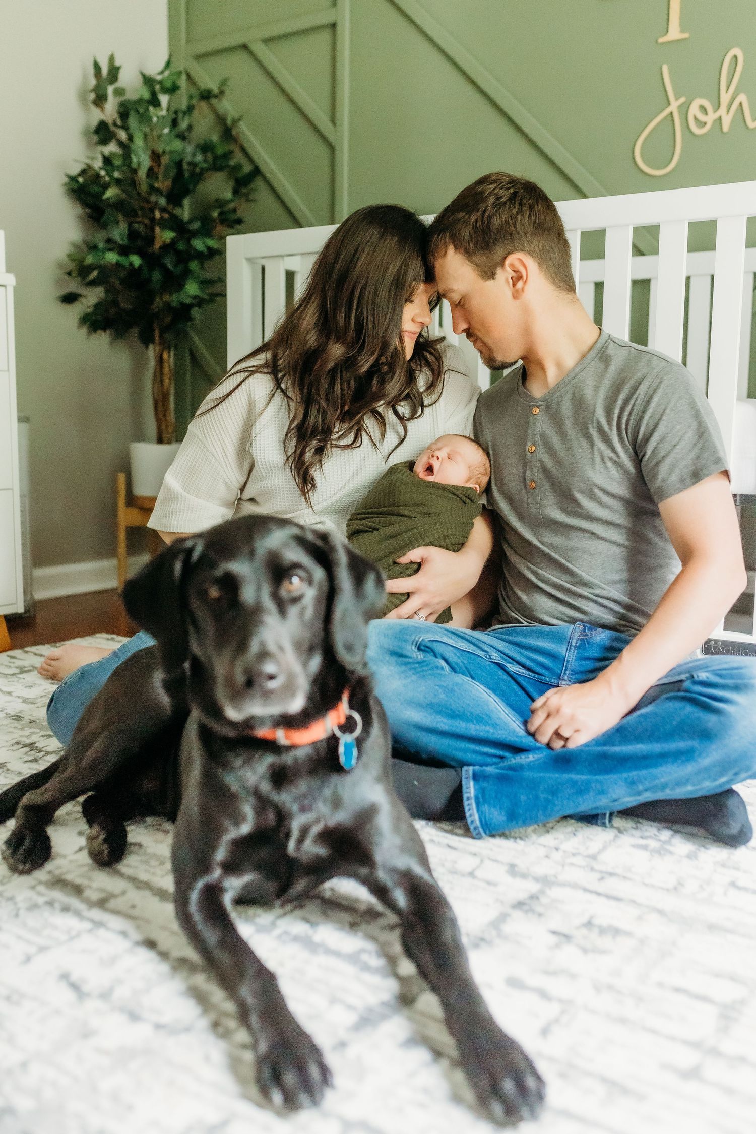 A couple shares an intimate moment with their newborn while their black dog lounges in front.
