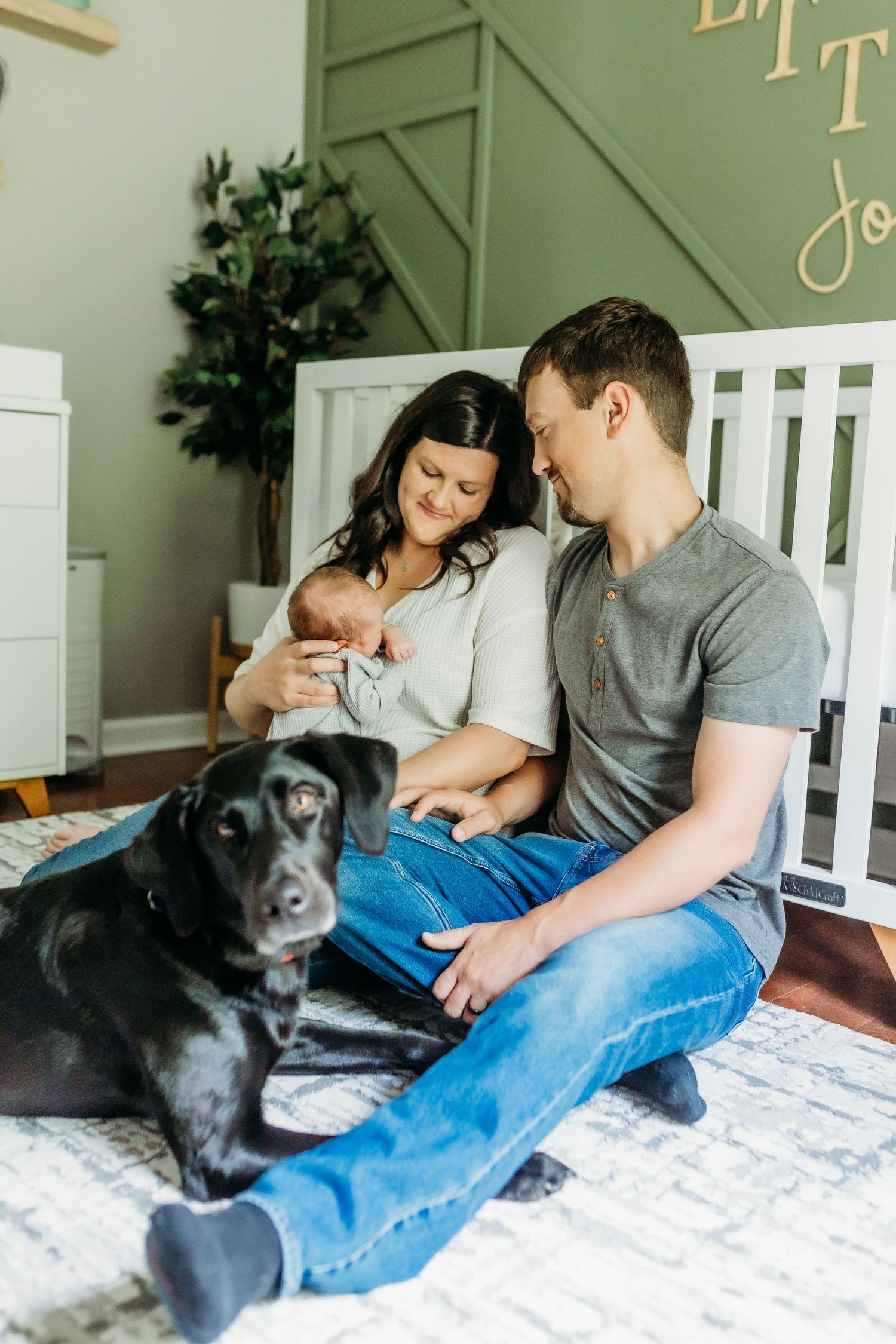 A family sits on the floor with their black dog while cradling their newborn in a nursery.