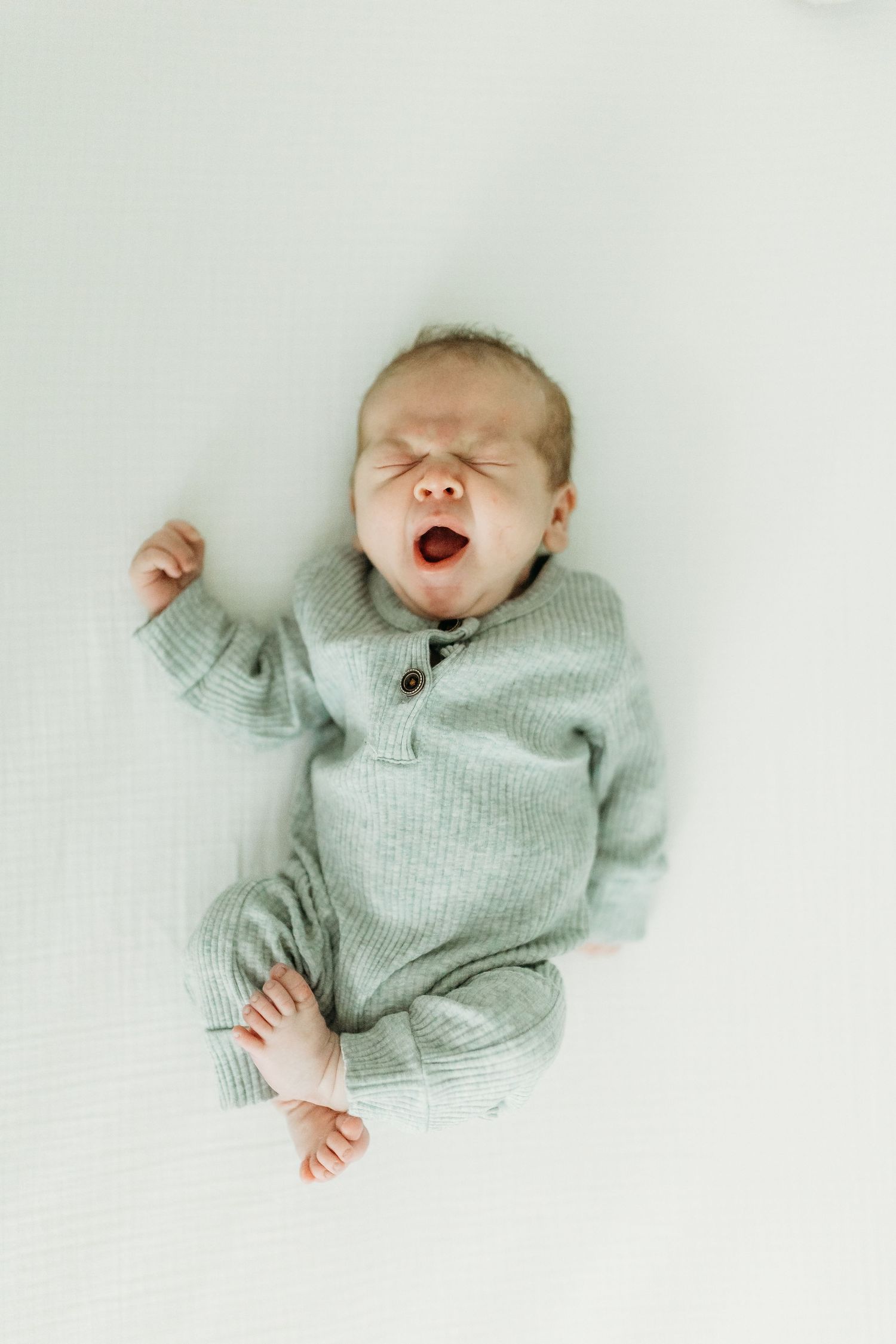 A yawning newborn baby wearing a light gray knit outfit lies on white fabric.