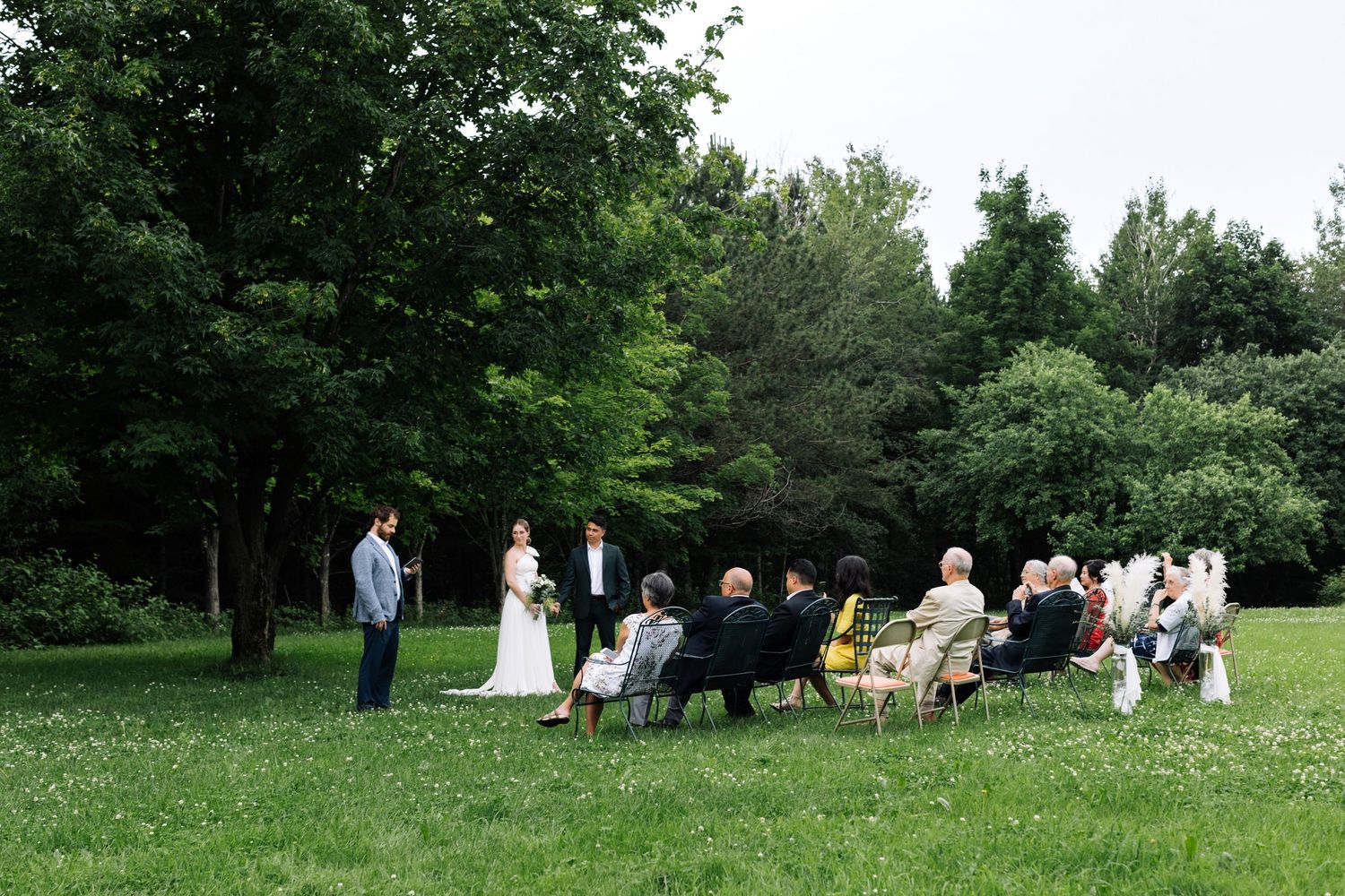 An intimate outdoor wedding ceremony with guests seated in chairs on a grassy lawn surrounded by trees.