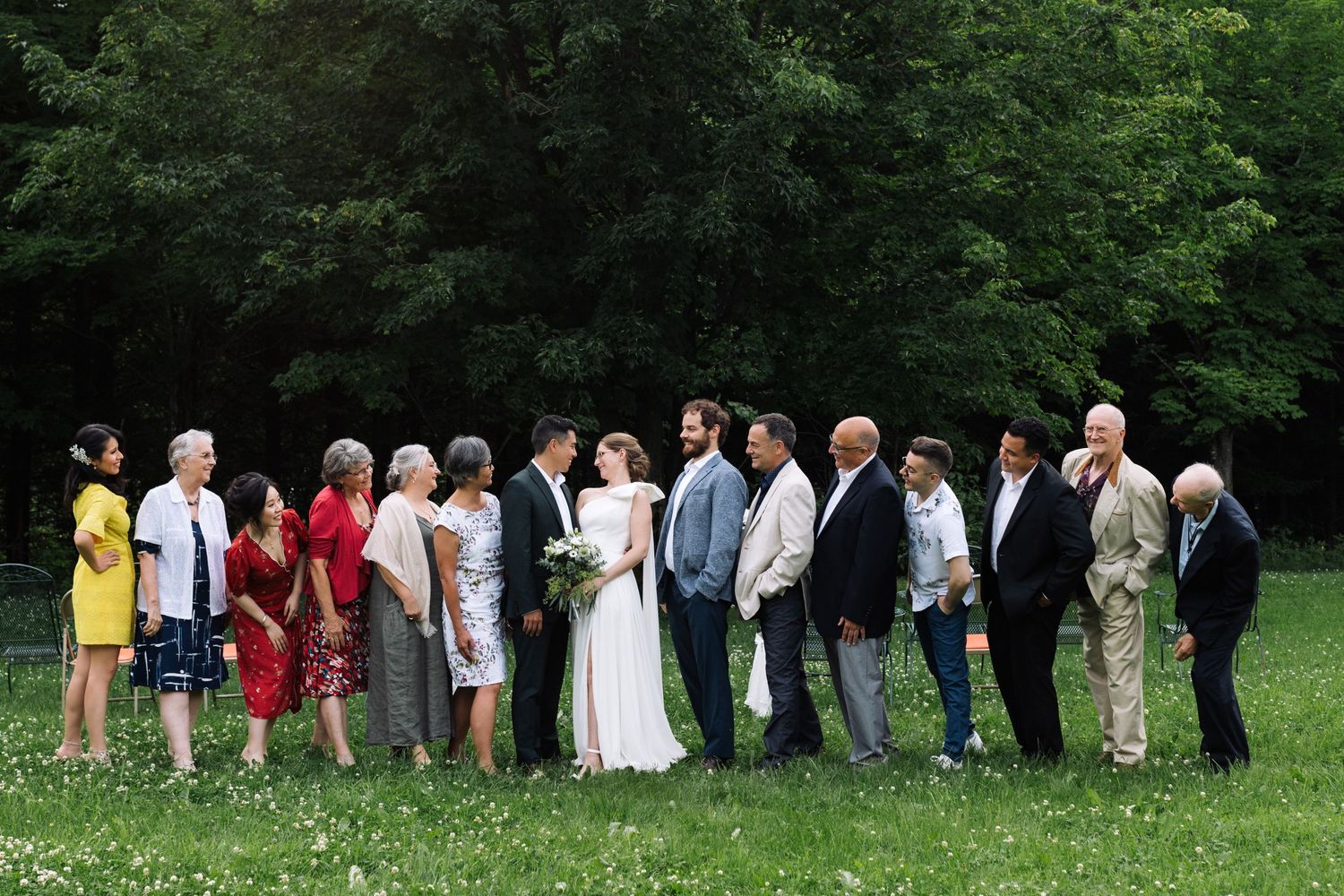 Wedding guests and the wedding party lined up for a group photo on grassy field with trees in background.