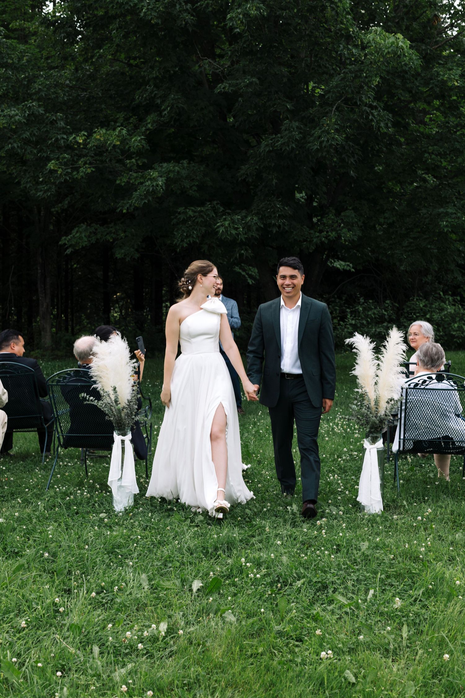 A wedding ceremony in a garden setting with white pampas grass decorations and guests seated on chairs.