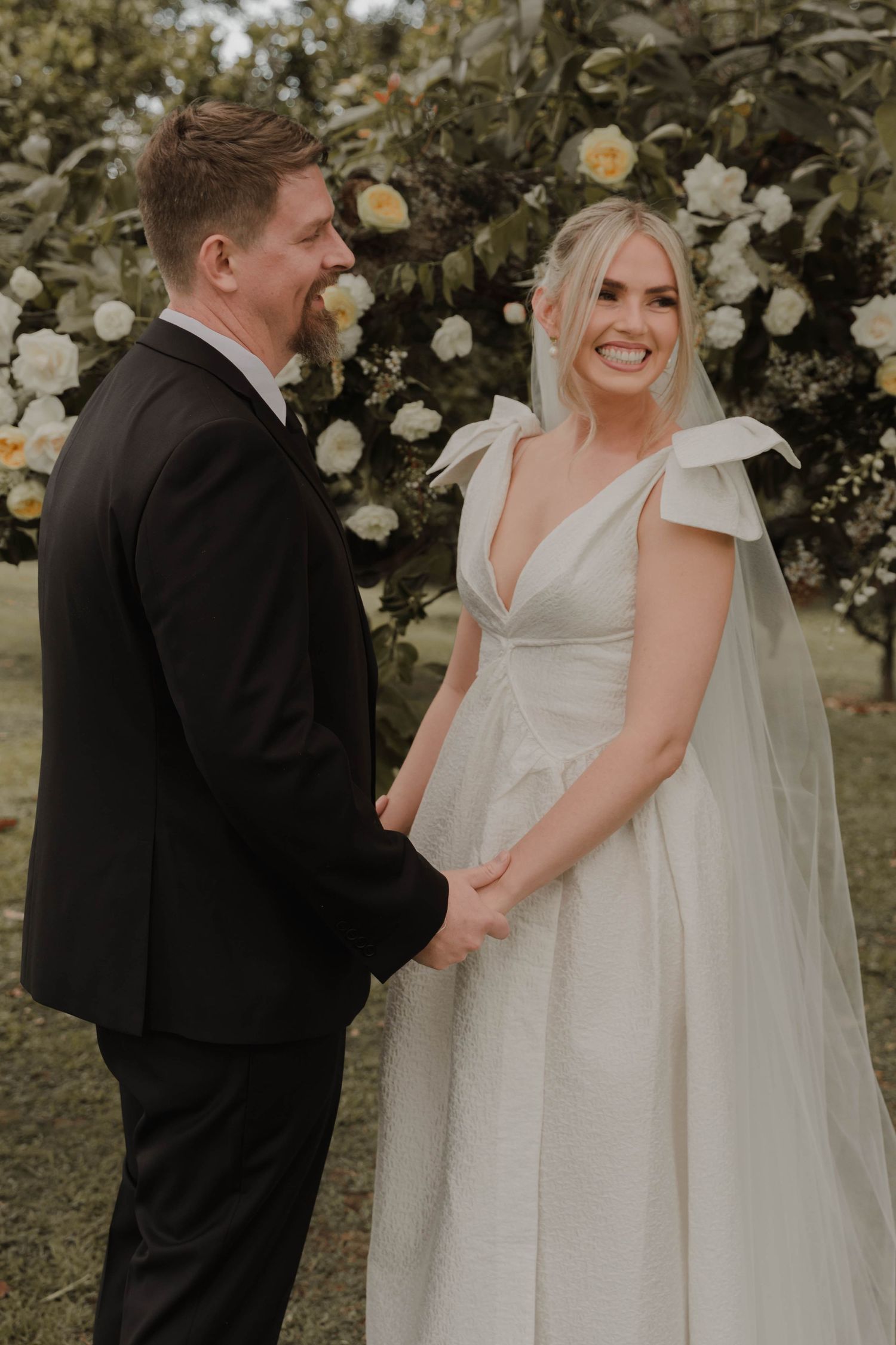 Joyful wedding couple sharing a moment in front of blooming white roses in an outdoor garden setting.