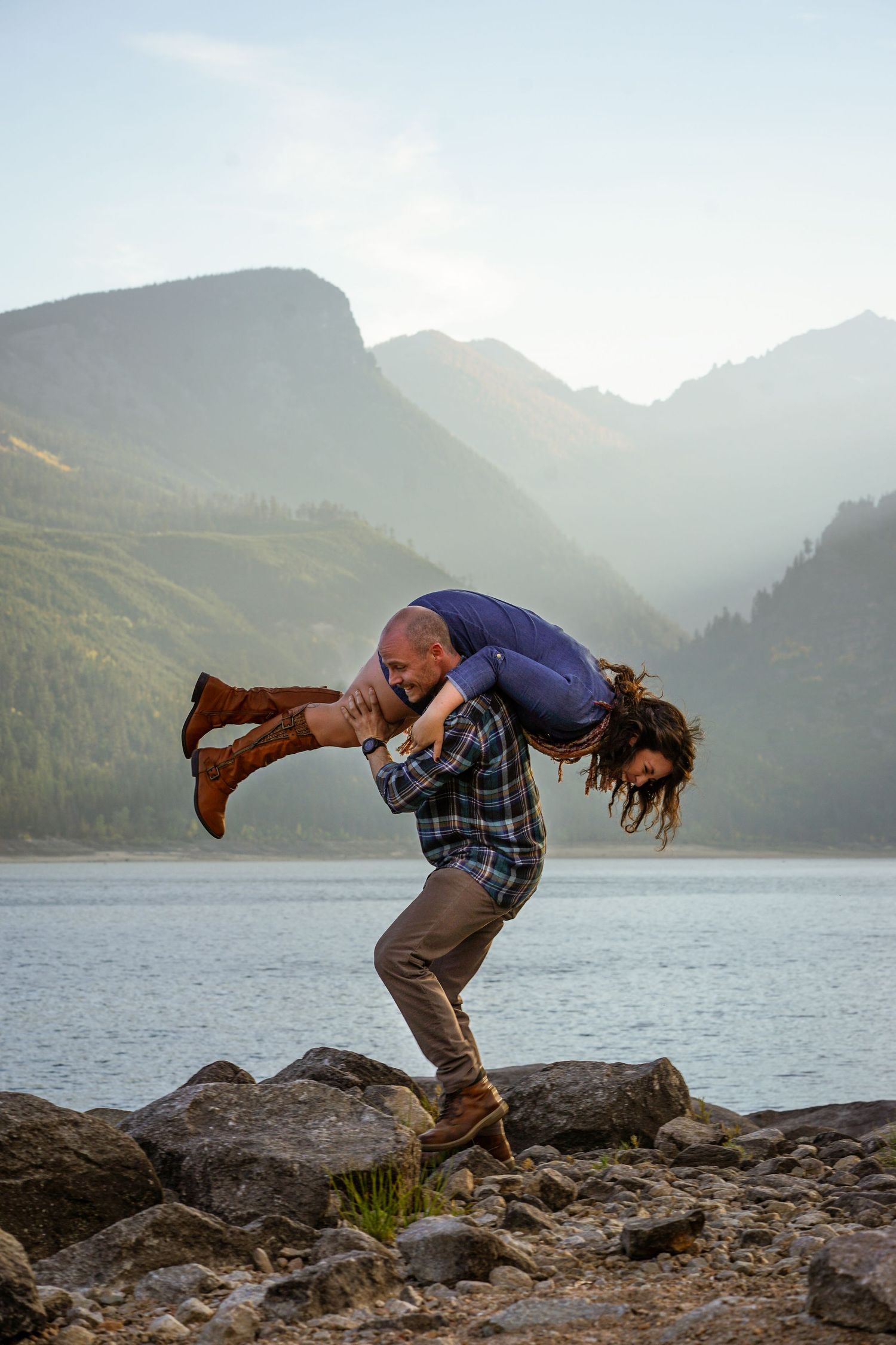 Playful couple poses dramatically on rocks with mountain lake backdrop.