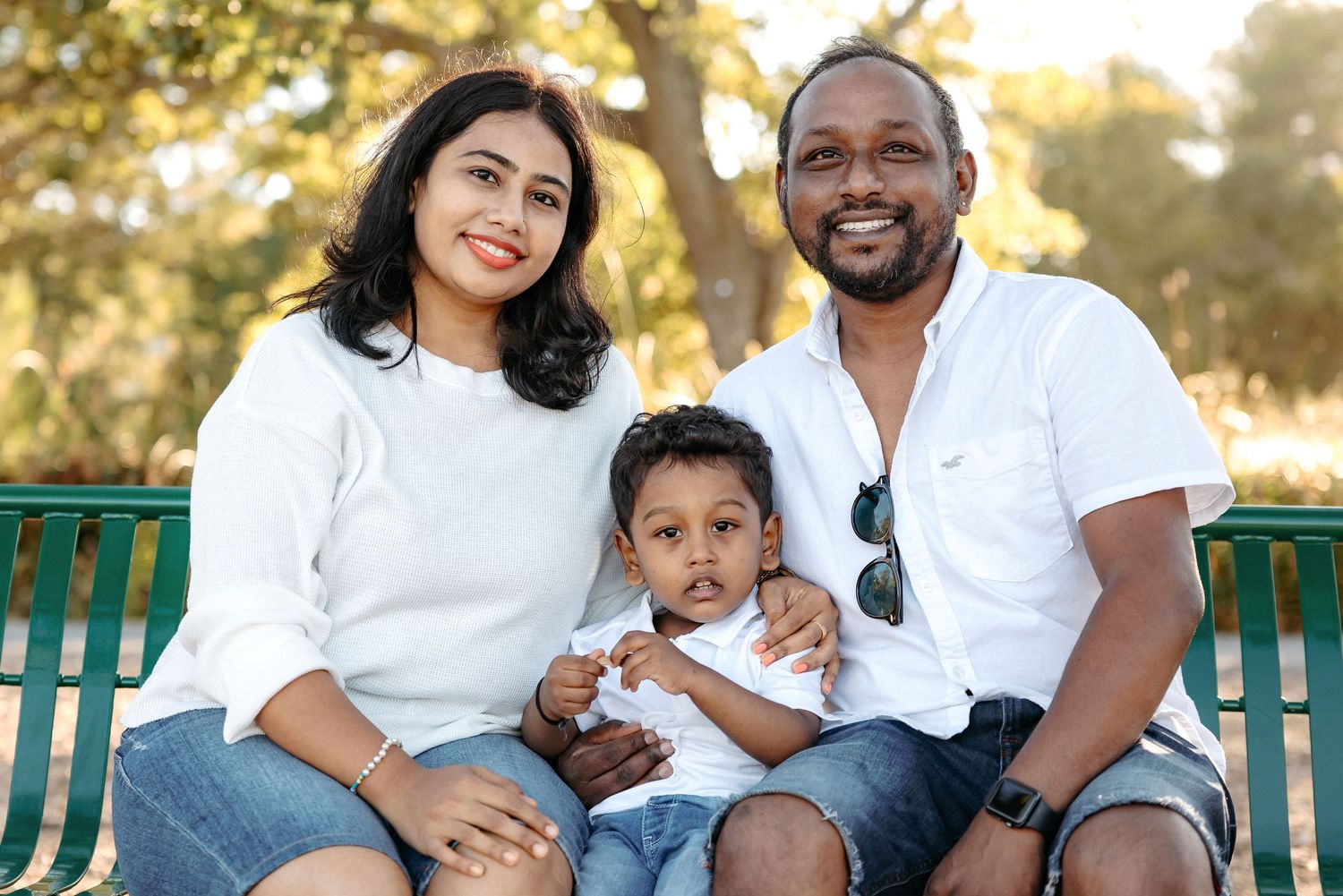 Family sitting together on a green park bench during a sunny outdoor photo session.