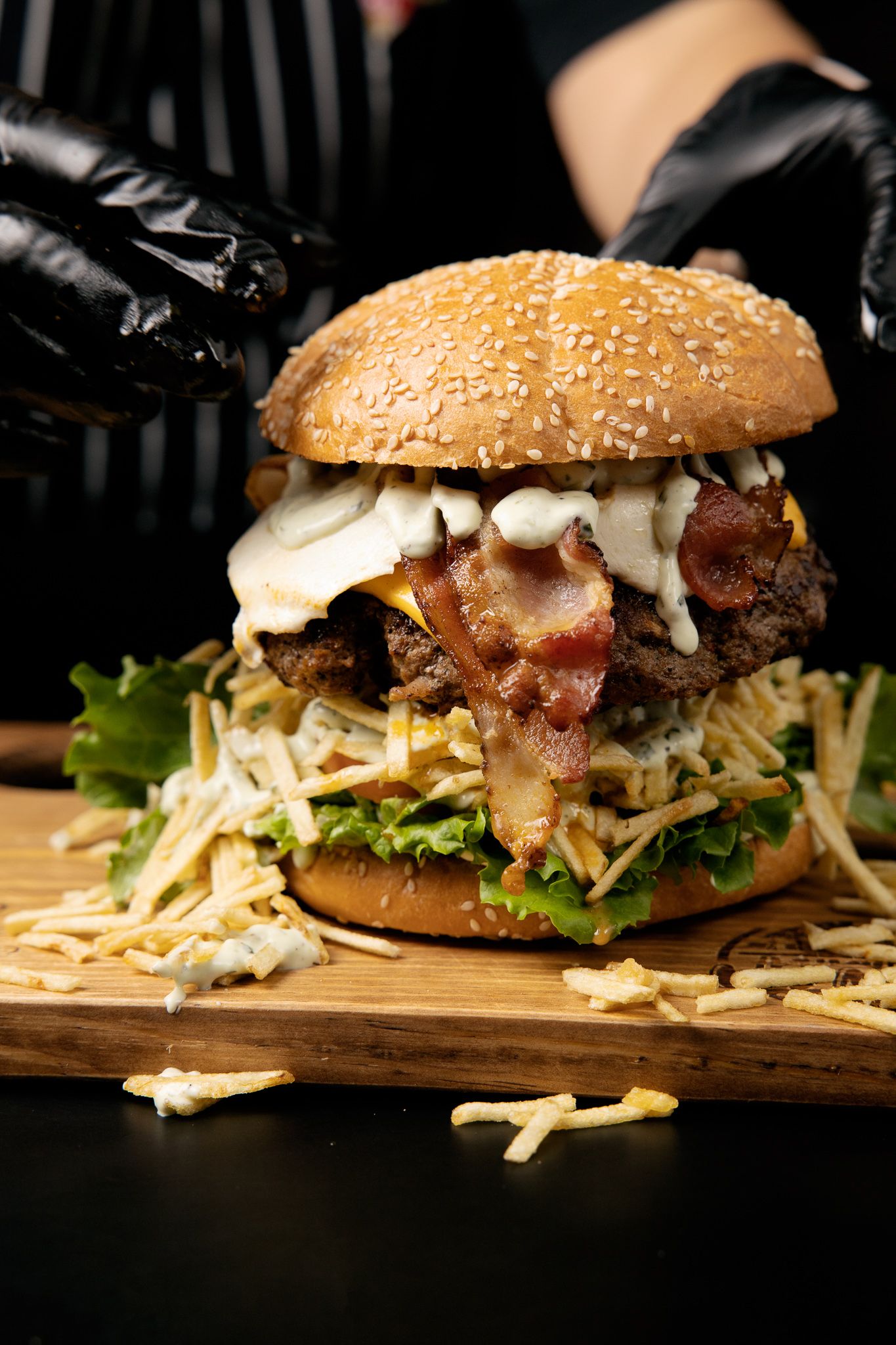 A towering gourmet burger with bacon, melted cheese and shredded lettuce on a wooden board.