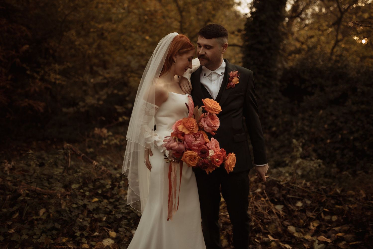A romantic autumn wedding photo featuring a bride in white holding vibrant orange flowers next to her groom.