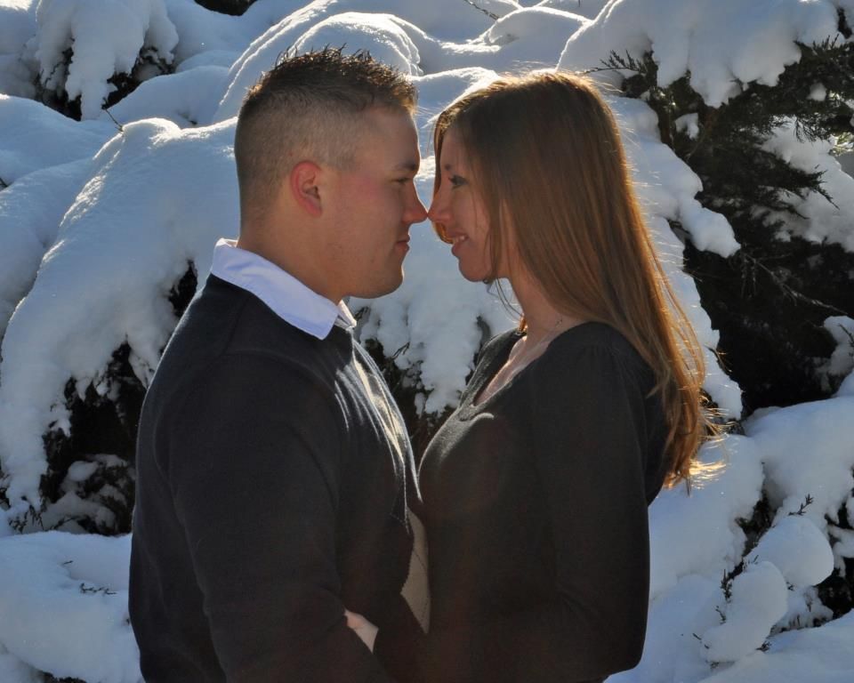 A romantic winter engagement photo with snow-covered evergreens in the background.