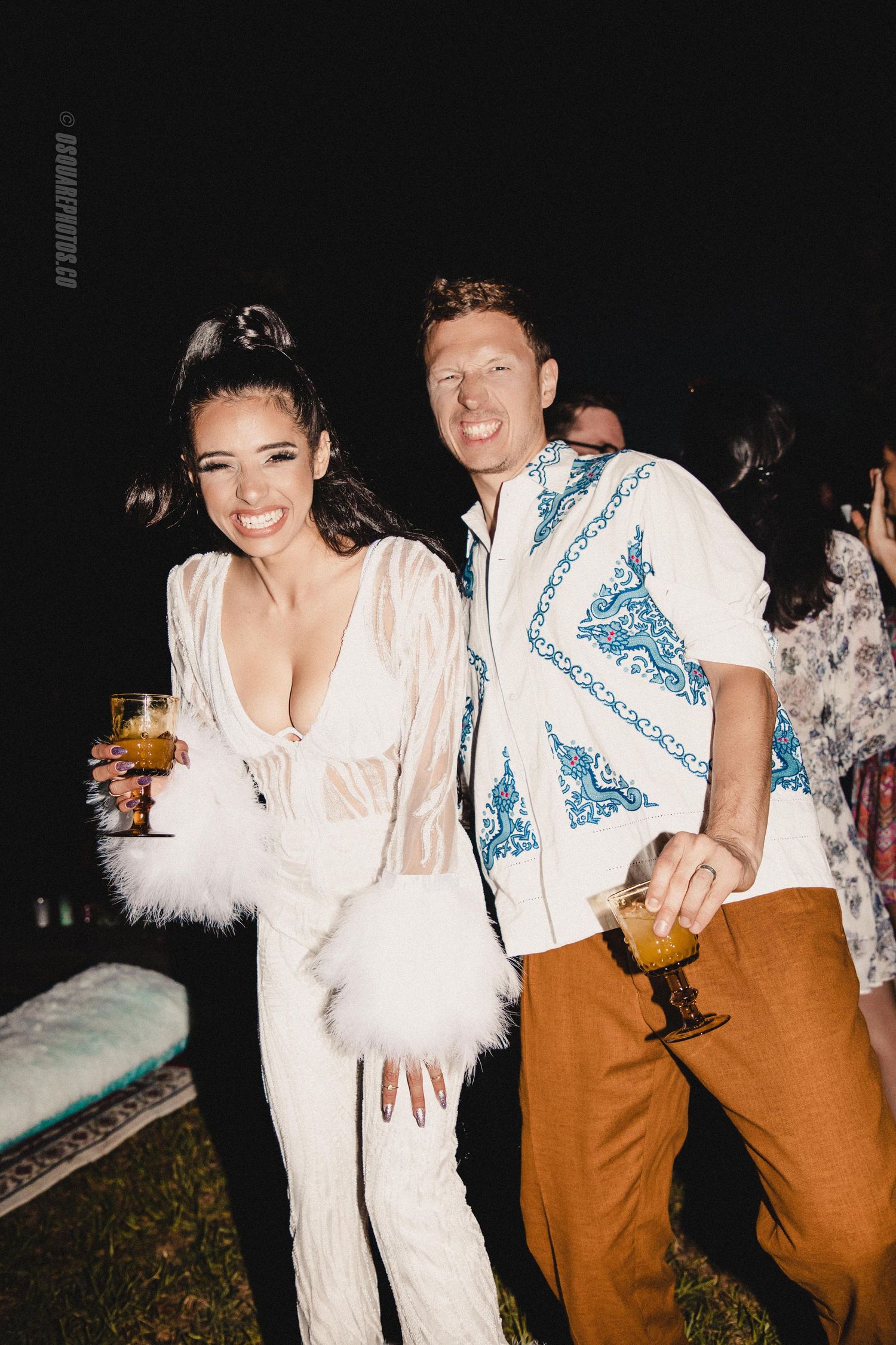 Partygoers in white and blue outfits smile while holding drinks at an outdoor gathering.