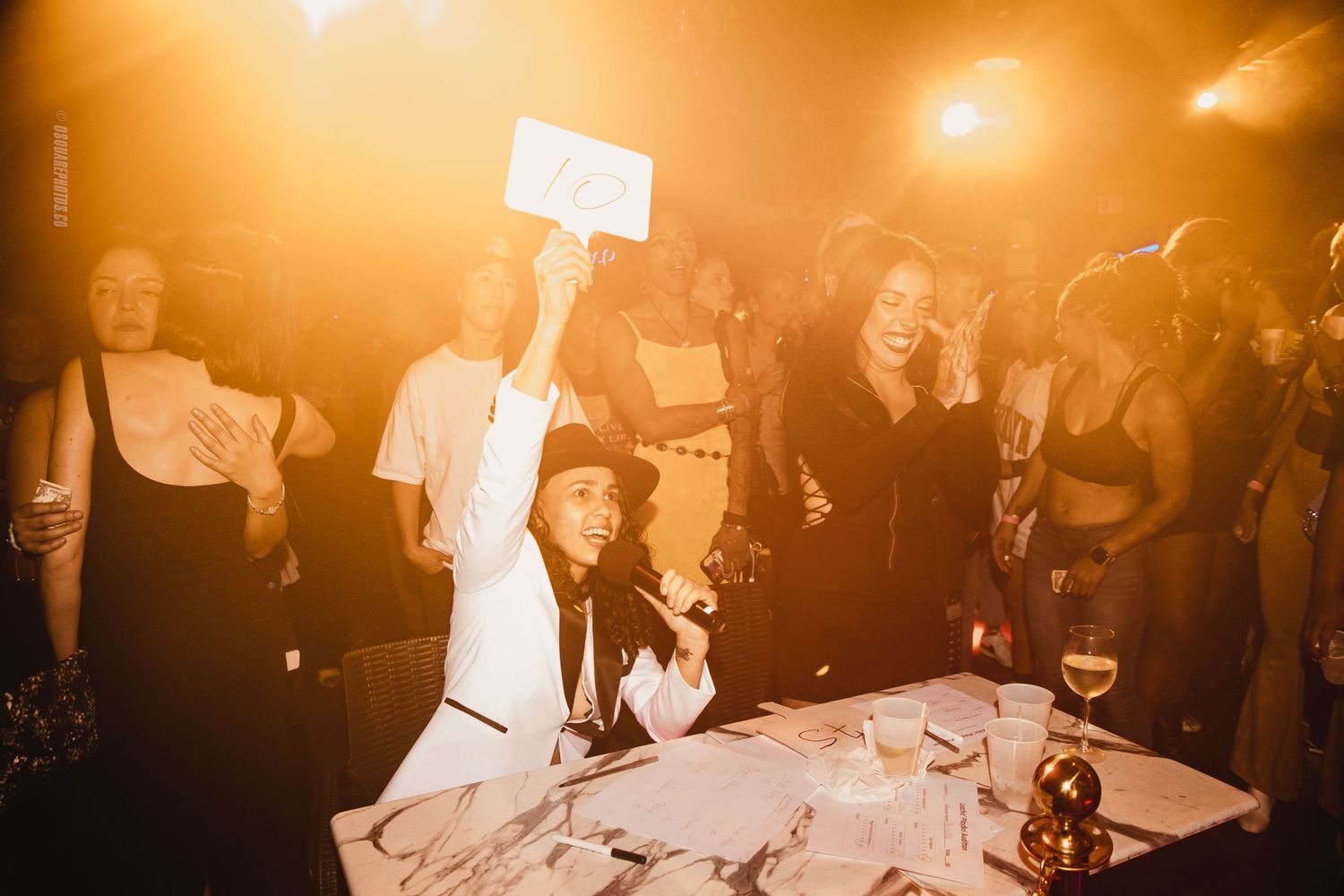 Lively party scene with people gathered around a table in warm orange lighting.