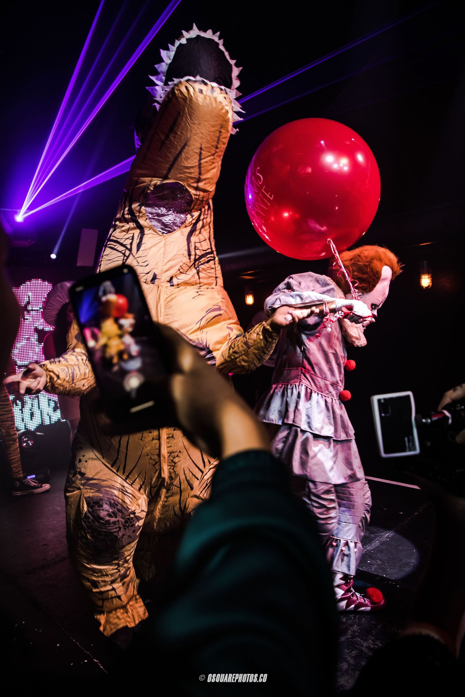 Inflatable dinosaur costume performer holding red balloon on stage with purple laser lights.