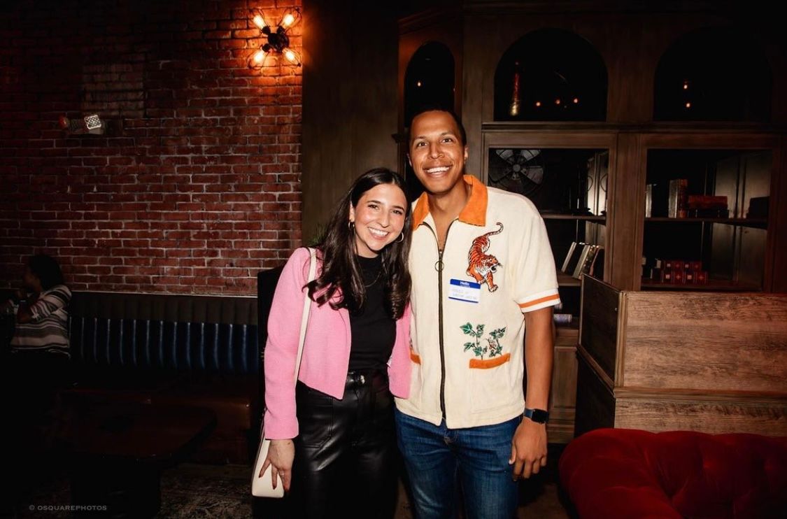 Two people posing together against a brick wall in a dimly lit venue with warm lighting.