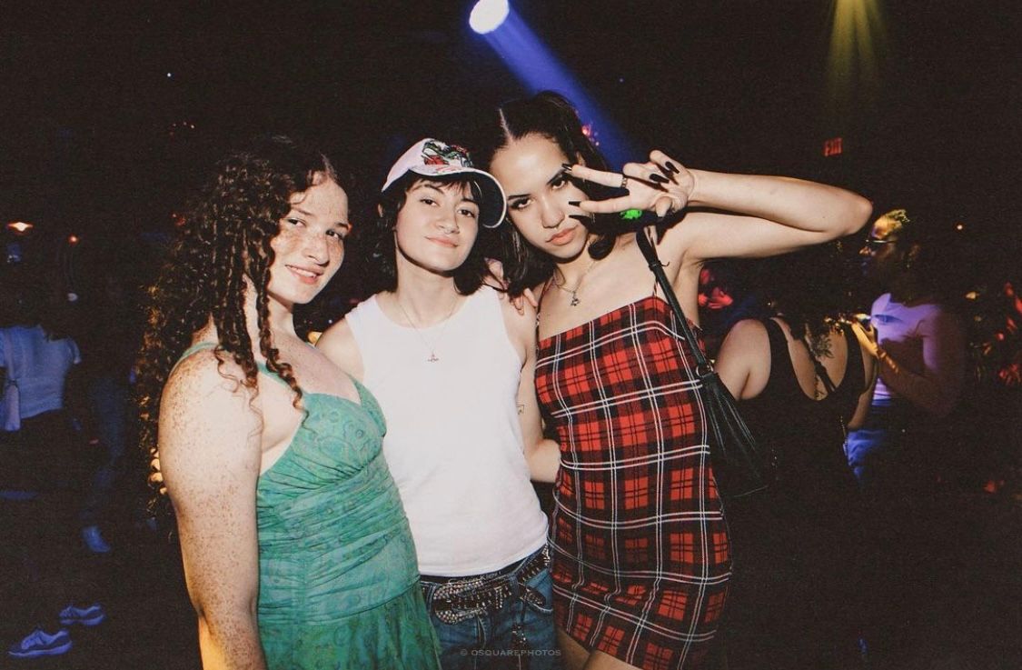 Three friends pose together at a nightclub, wearing colorful dresses and casual attire in a festive party atmosphere.