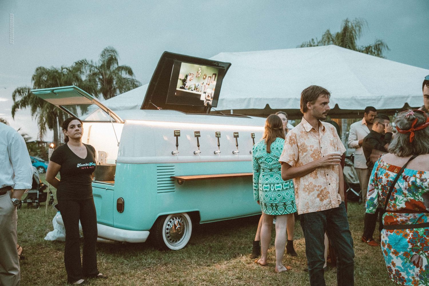 Classic blue and white VW van serves as mobile bar at outdoor festival with palm trees.
