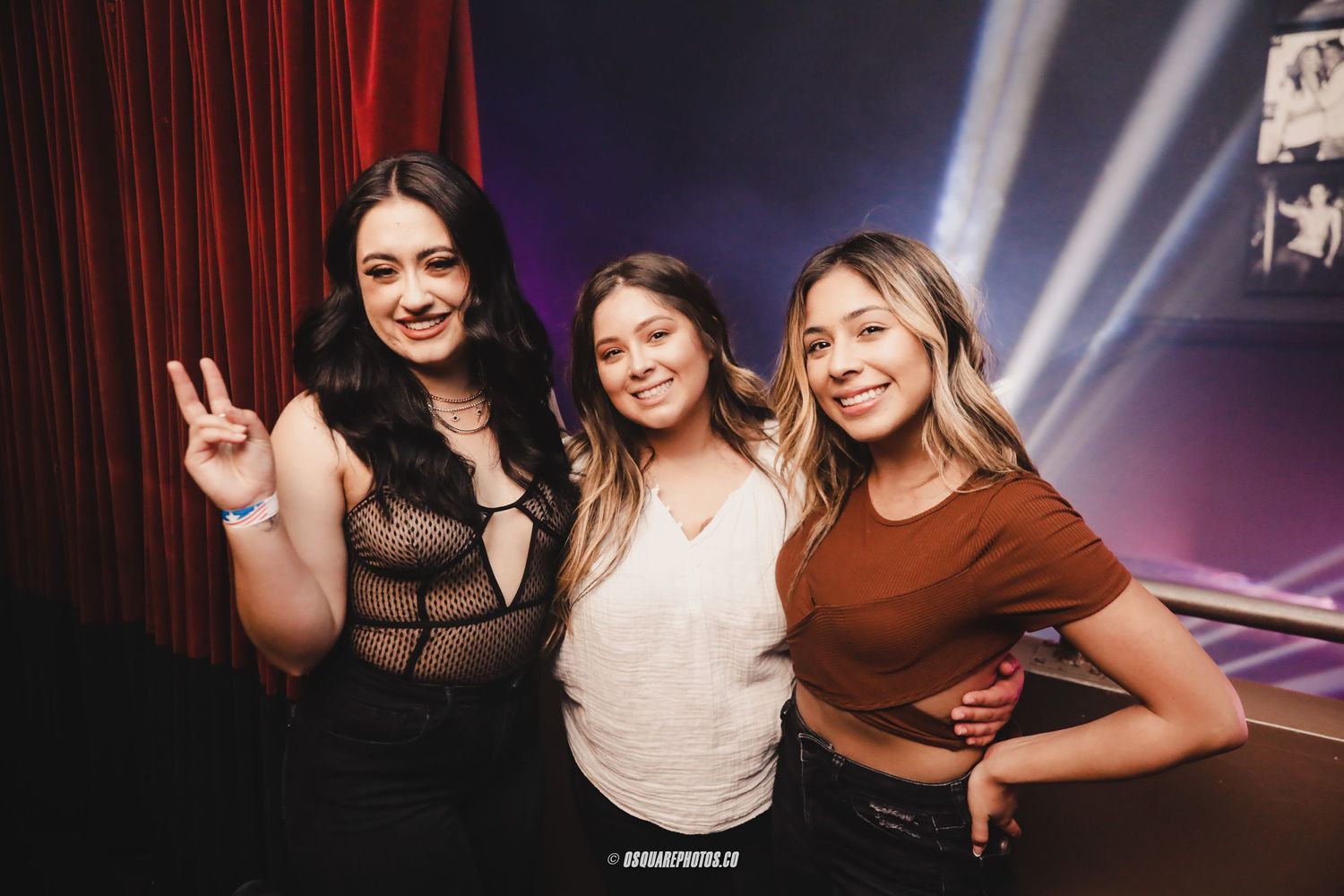 Three friends pose together at a nightclub event with dramatic lighting and red curtains in the background.