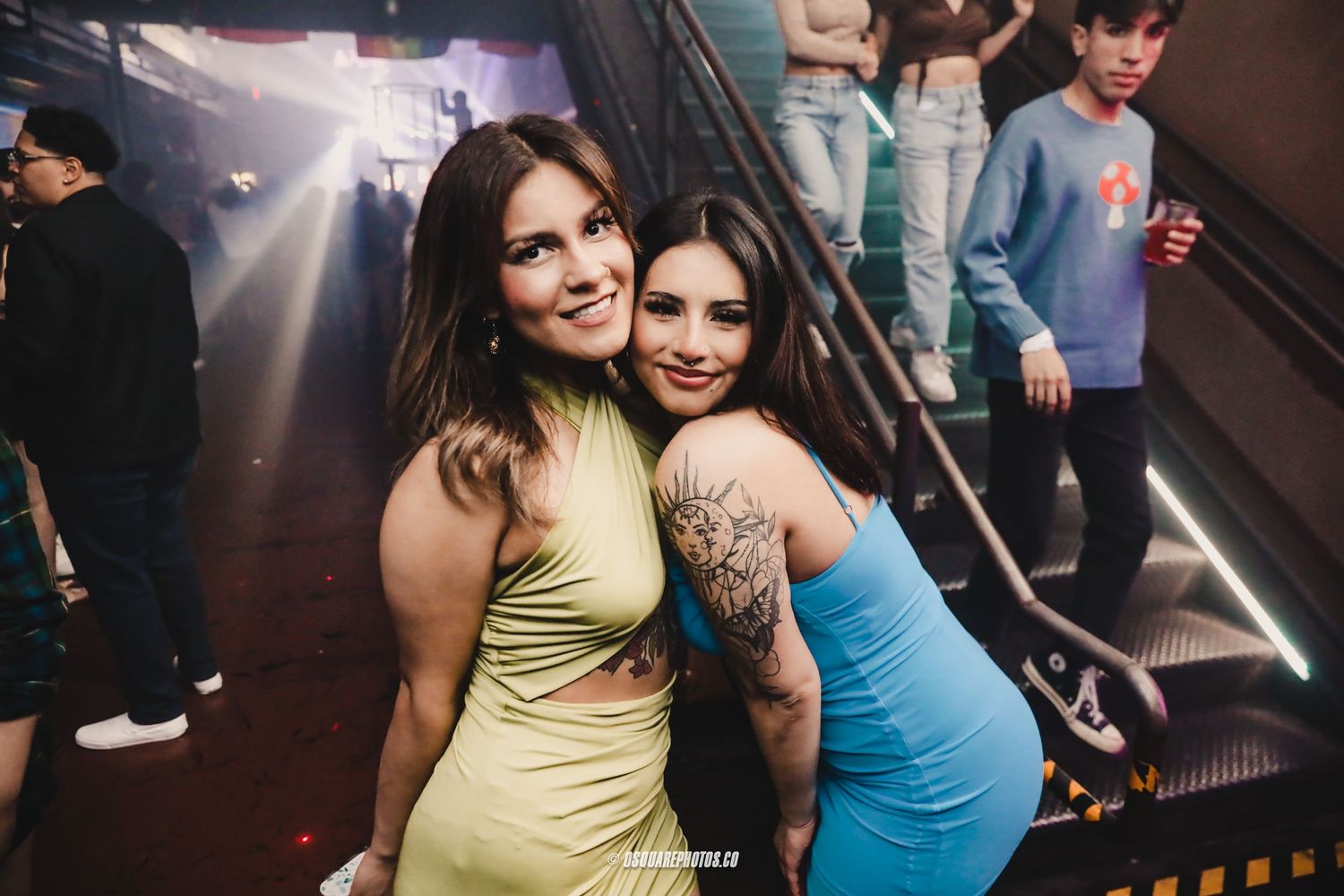 Two partygoers in colorful dresses pose together on a staircase at a club event.