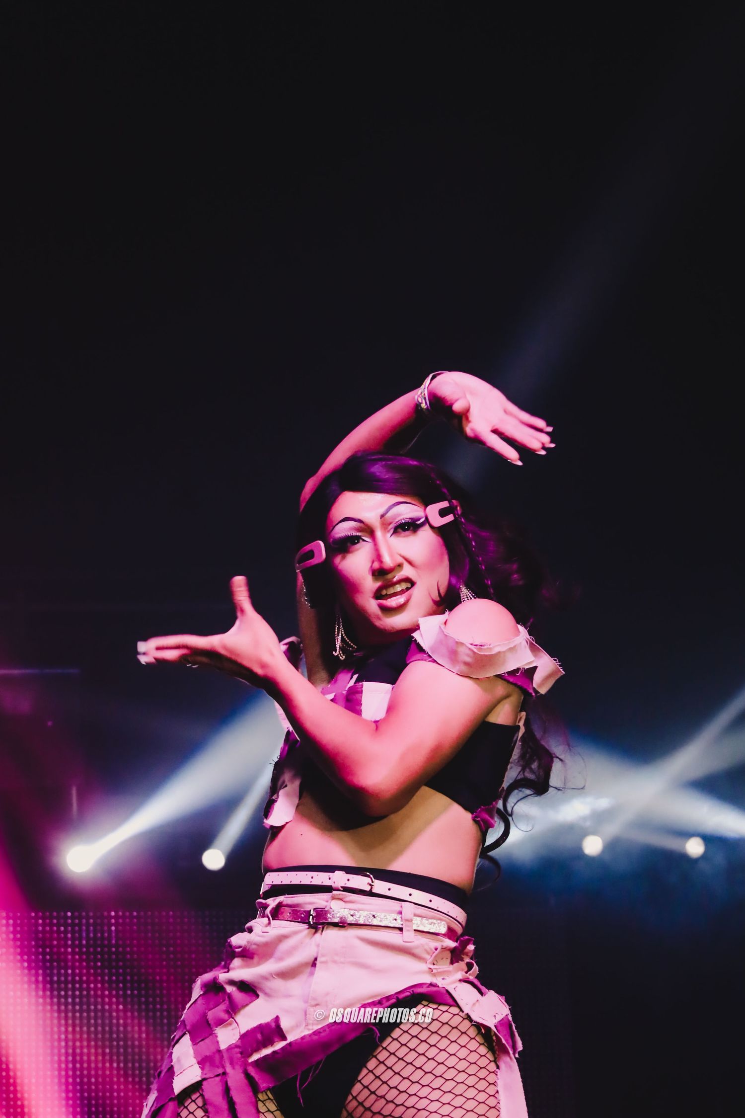 Performer in pink lighting strikes dramatic dance pose on stage with spotlight effects.