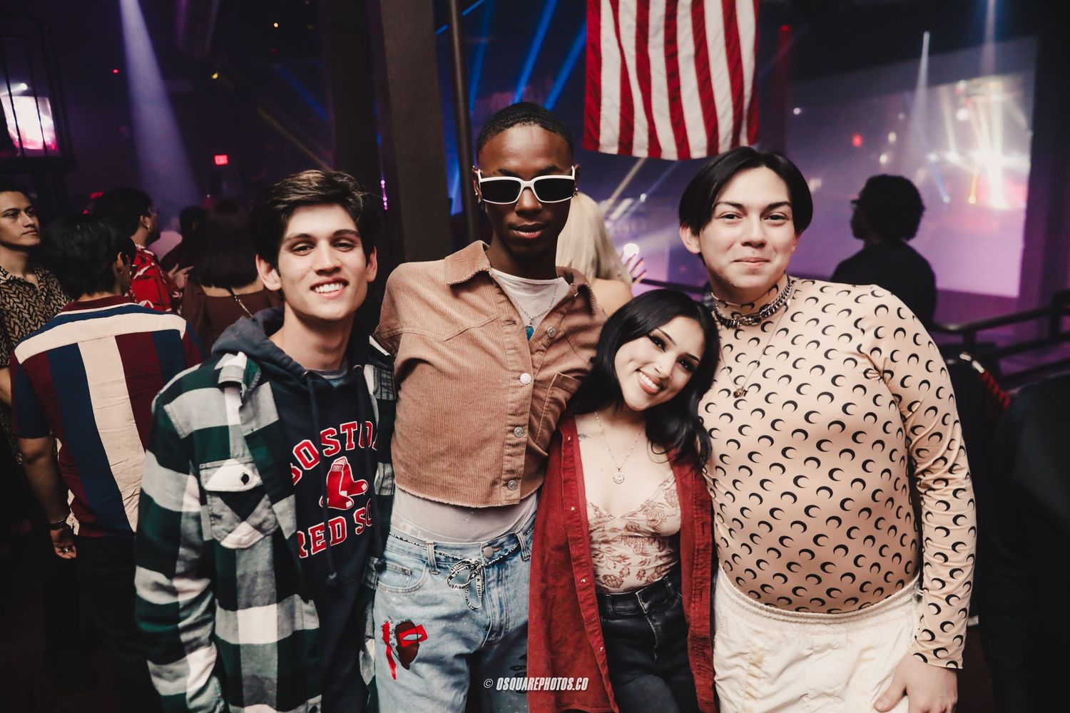 Diverse group of friends posing together at a club with American flag visible in background.
