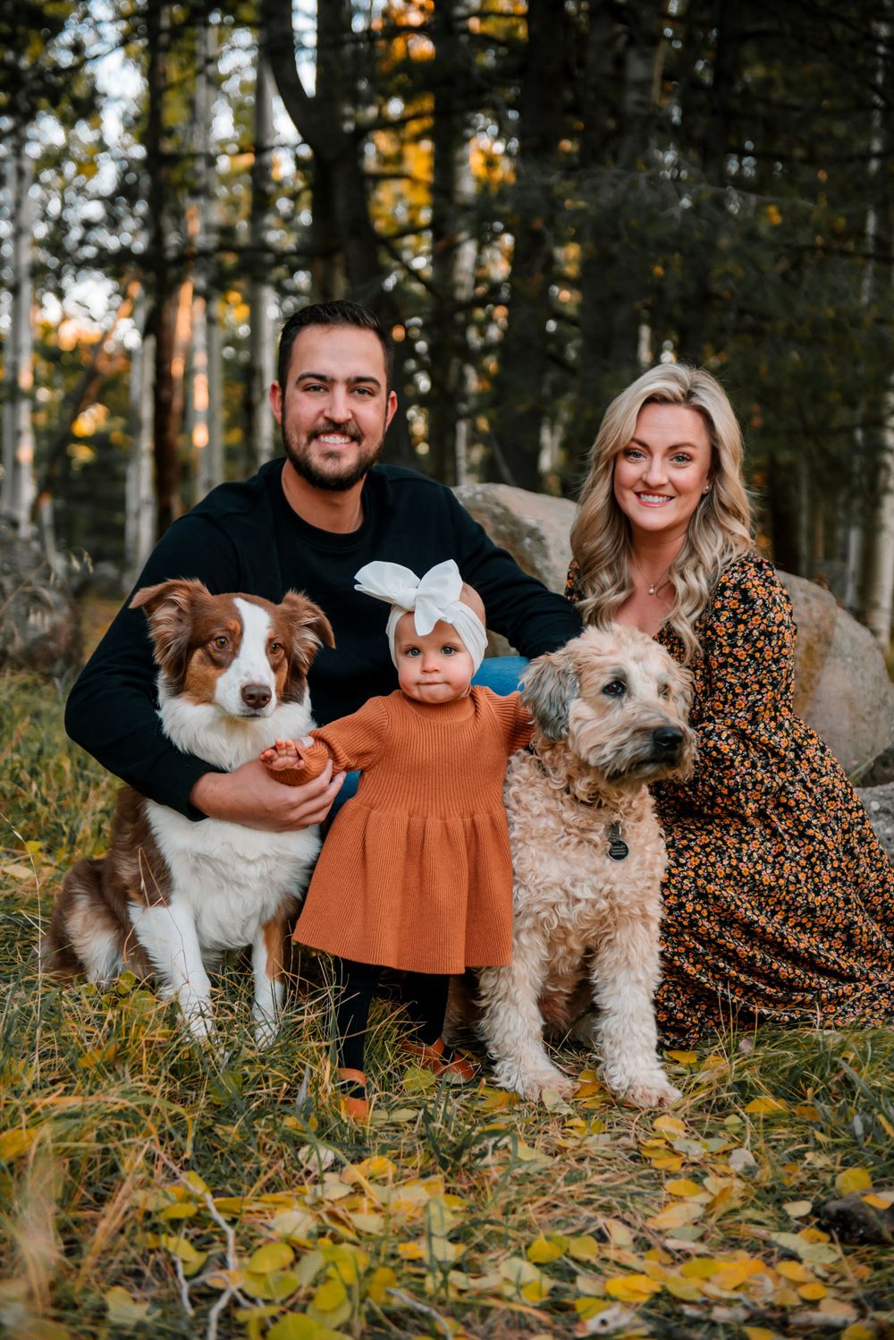 A family poses outdoors with their two dogs in an autumn forest setting.