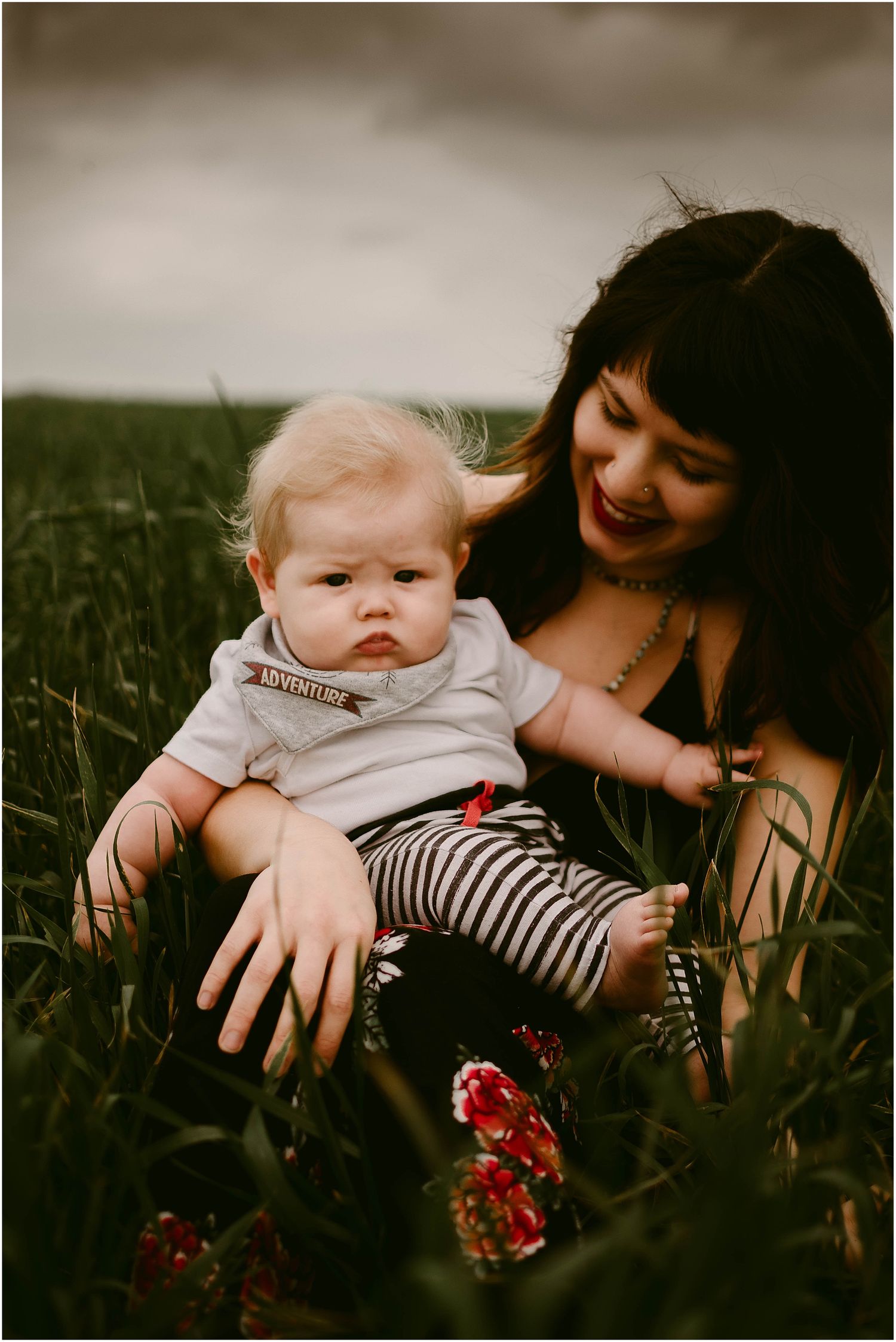 A baby in striped pants and white shirt sits in tall grass near red wildflowers.