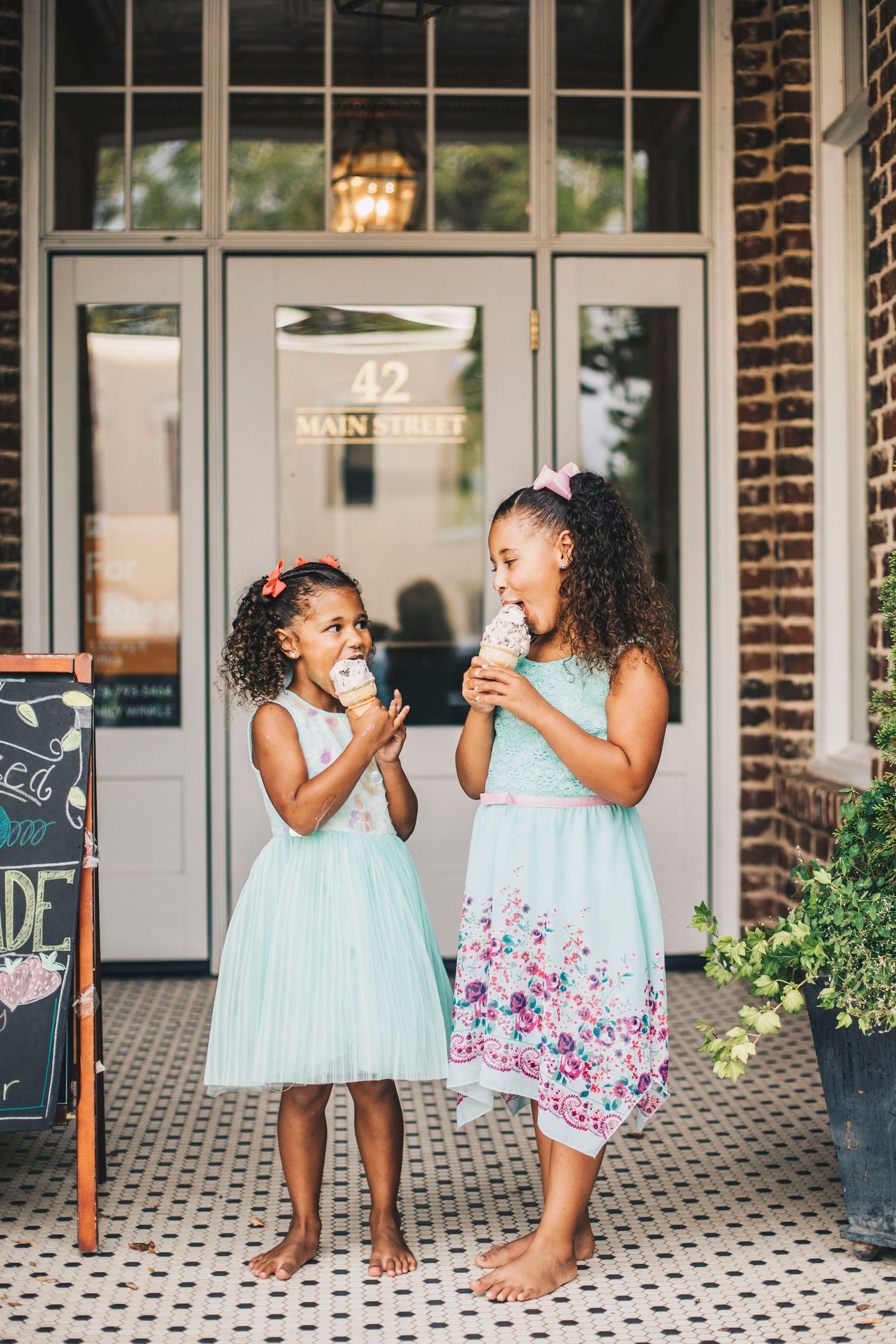 Two young girls in matching mint dresses eating ice cream cones outside a shop.