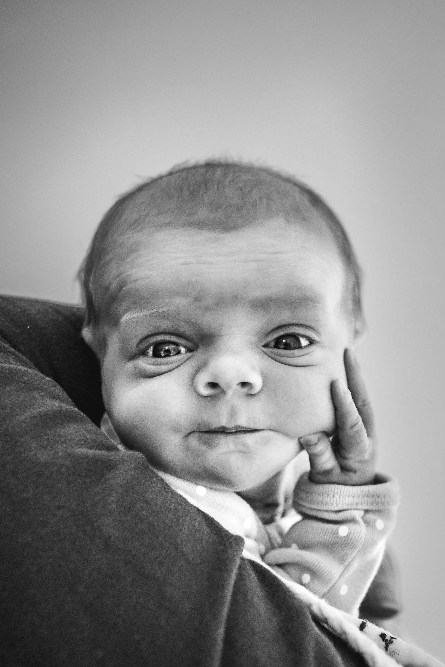 Very wide eyed and aware newborn baby looks at the camera with her hand on her cheek.