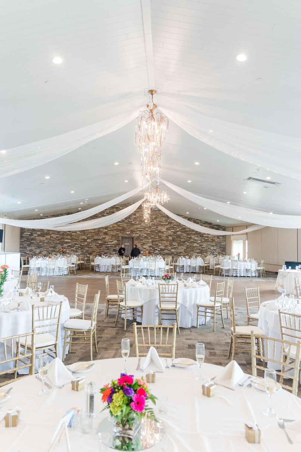 Wedding reception hall with white draped ceiling, chandeliers and elegant table settings.