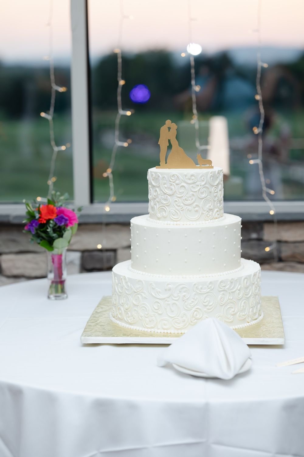 Elegant three-tier white wedding cake with lace details and gold topper against sunset window view.