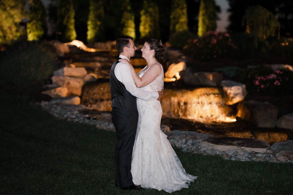 Romantic evening wedding portrait with couple embracing in front of lit stone wall.