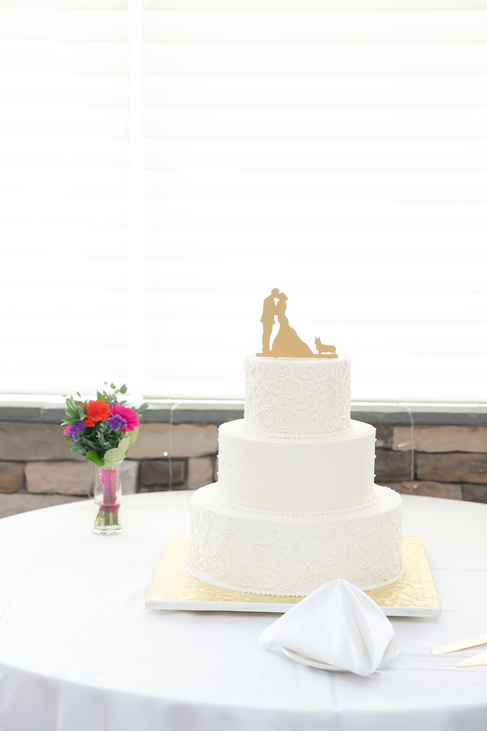 White tiered wedding cake with silhouette topper and pink floral accent on round table.