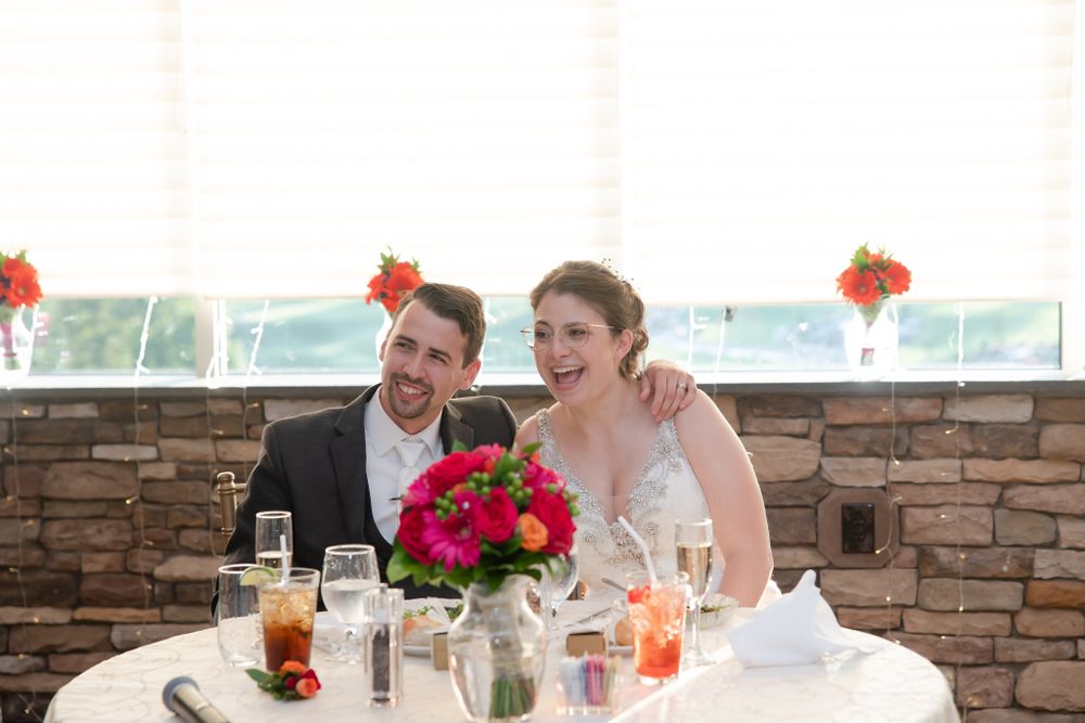 A couple shares a joyful moment at their wedding reception table with vibrant red floral centerpiece.