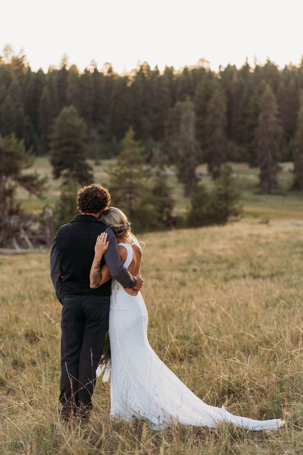 Newlywed couple gaze across mountain meadow with forest backdrop at sunset in wedding attire.
