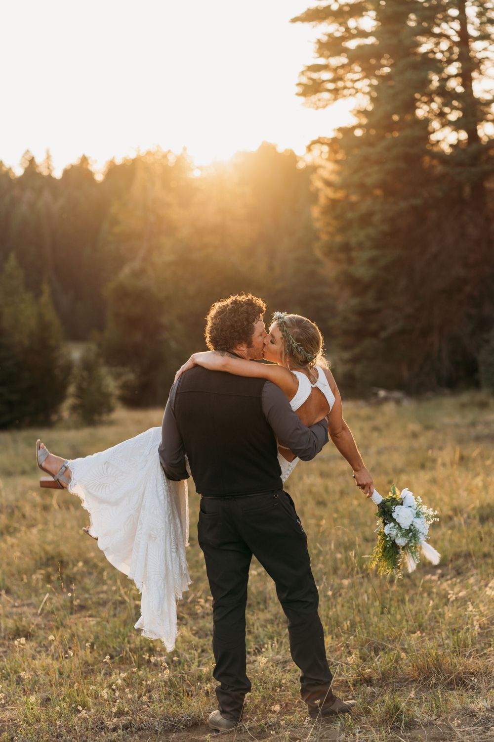 Groom carries bride in sunset meadow while holding white floral bouquet.