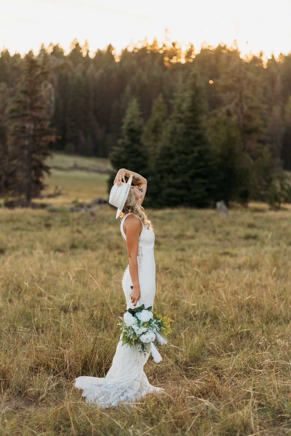 Bride in lace dress holds bouquet while standing in mountain meadow at sunset.