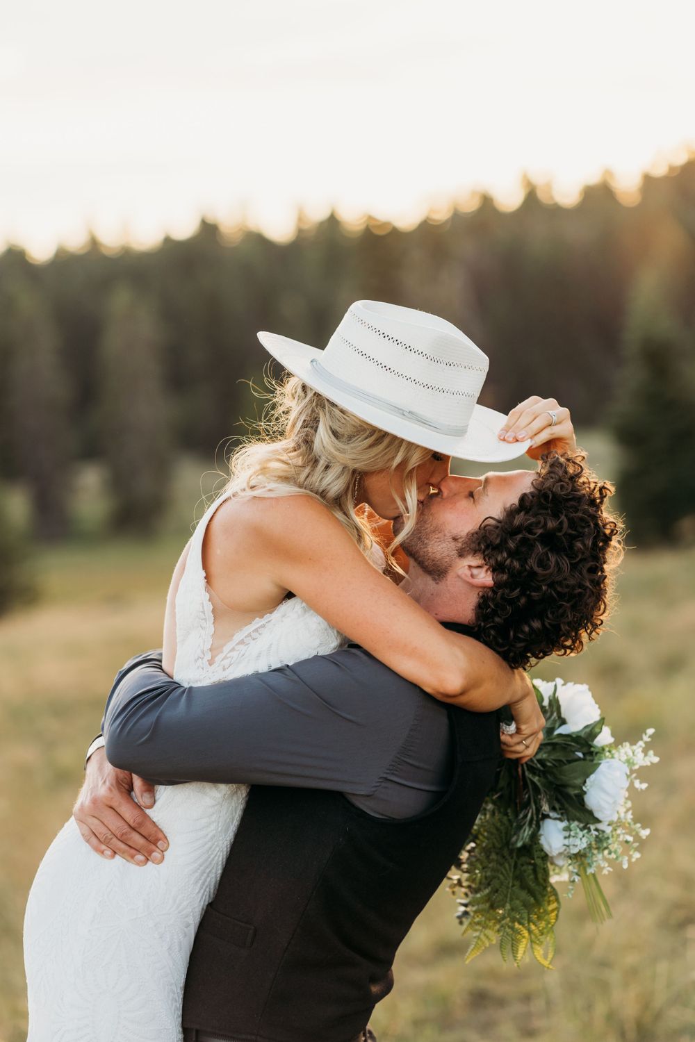Joyful wedding couple embraces in meadow with bride wearing cowboy hat.