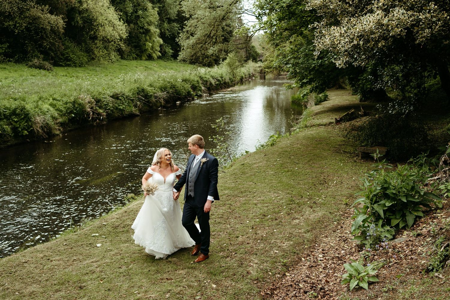 Newlyweds share romantic moments along tree-lined river path.