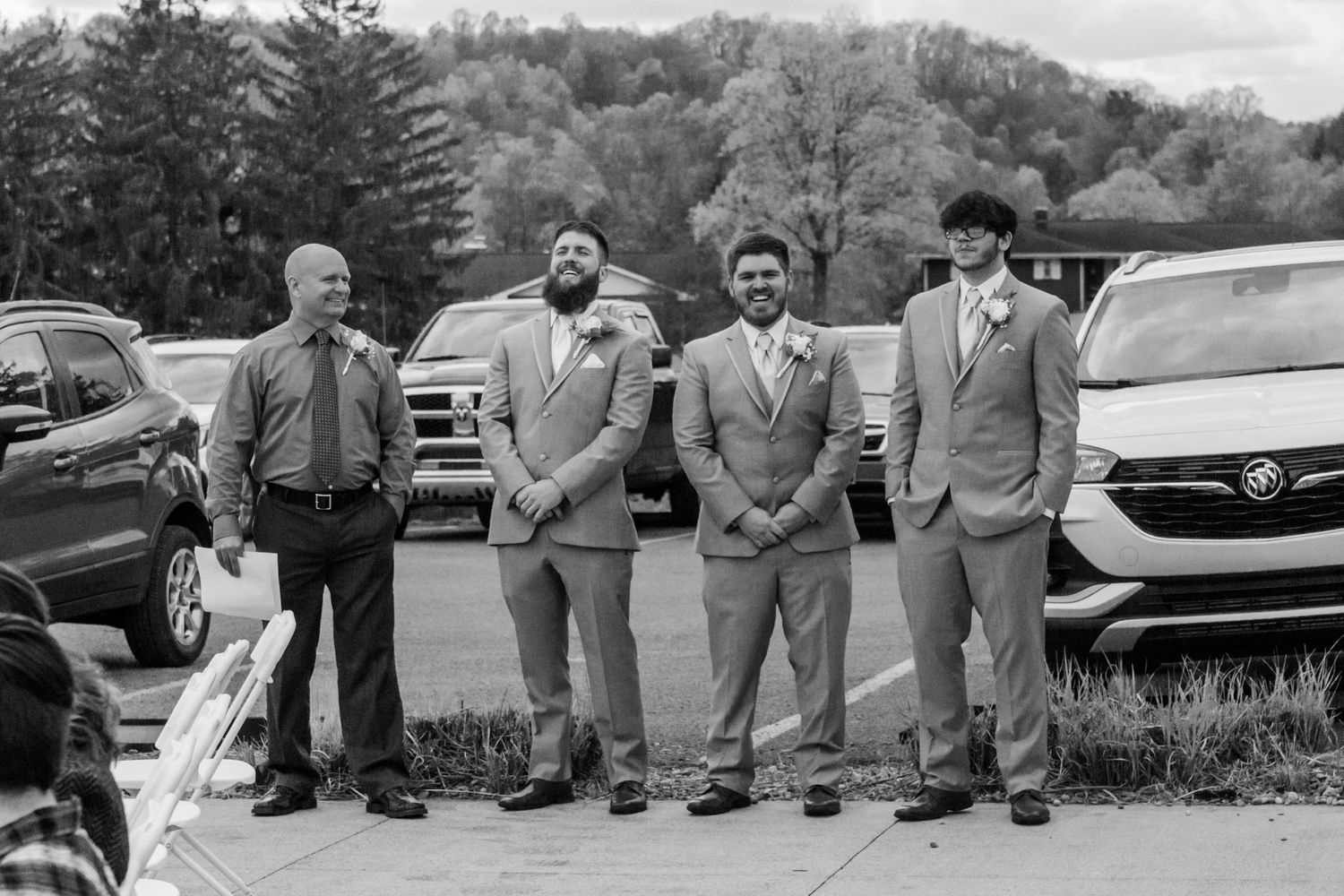 Wedding party members in gray suits sit casually on a wooden fence outdoors.