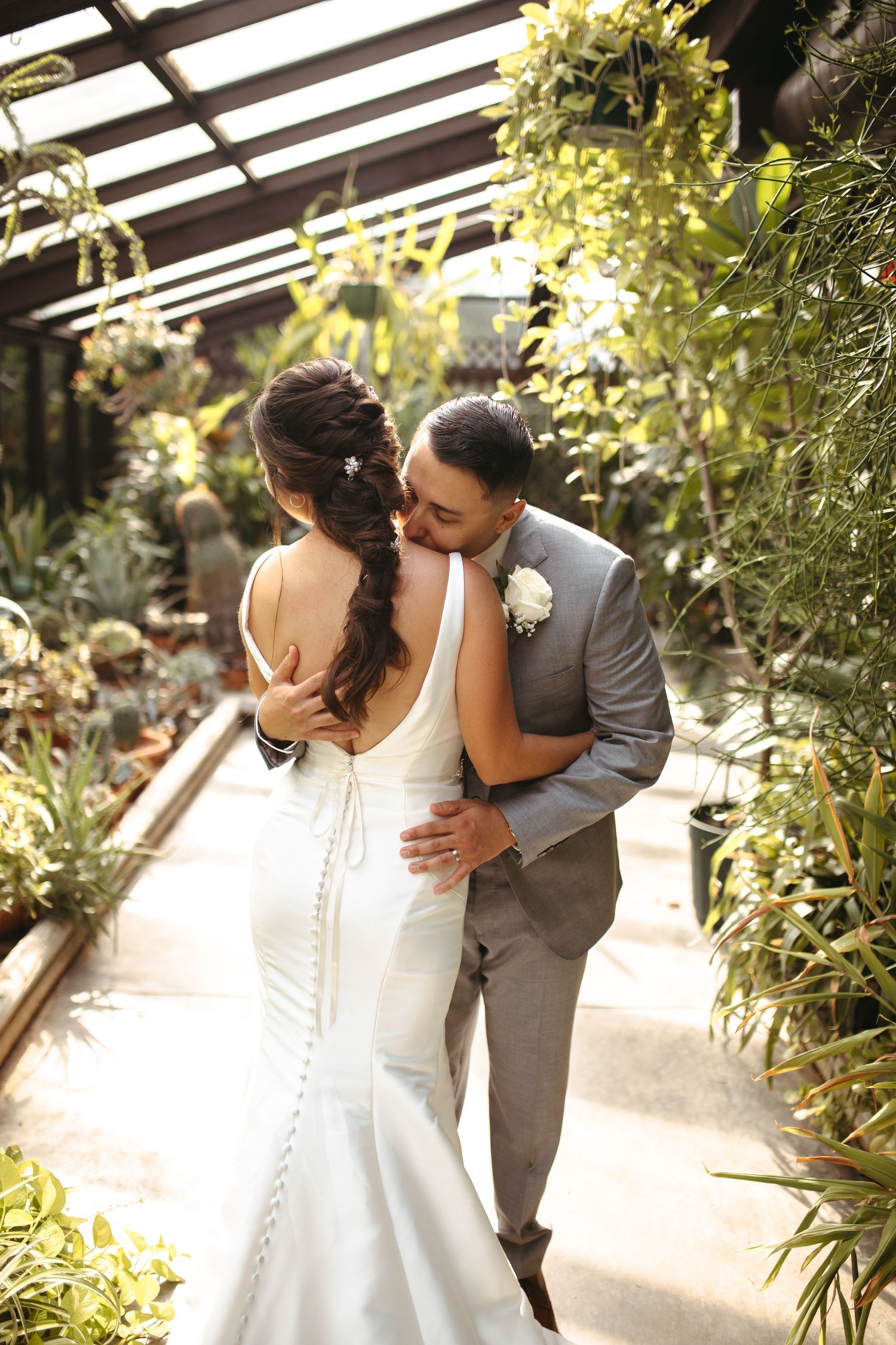 Wedding couple embracing in lush greenhouse with light streaming through glass ceiling.