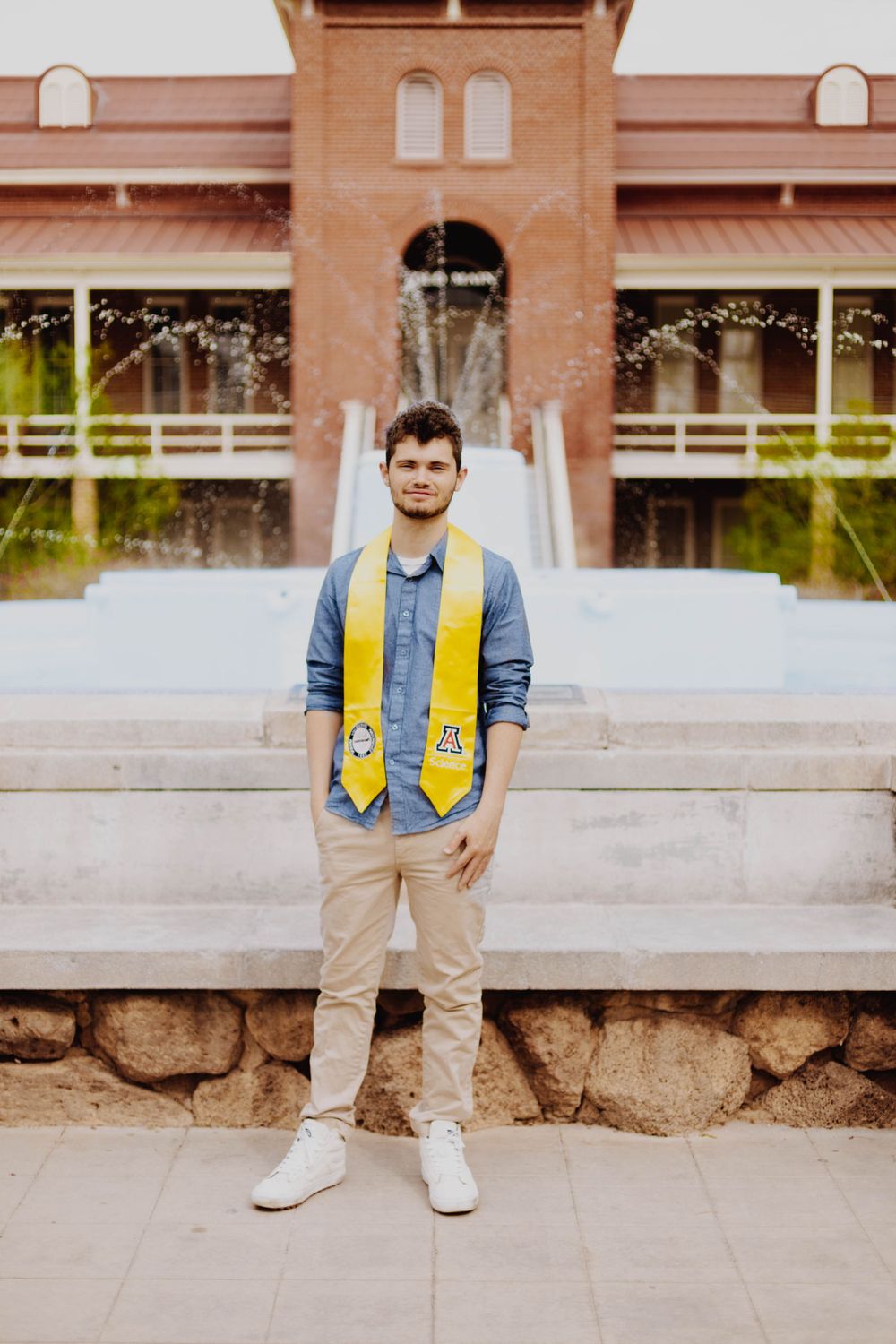 Graduate in denim and yellow stoles poses against brick building backdrop.